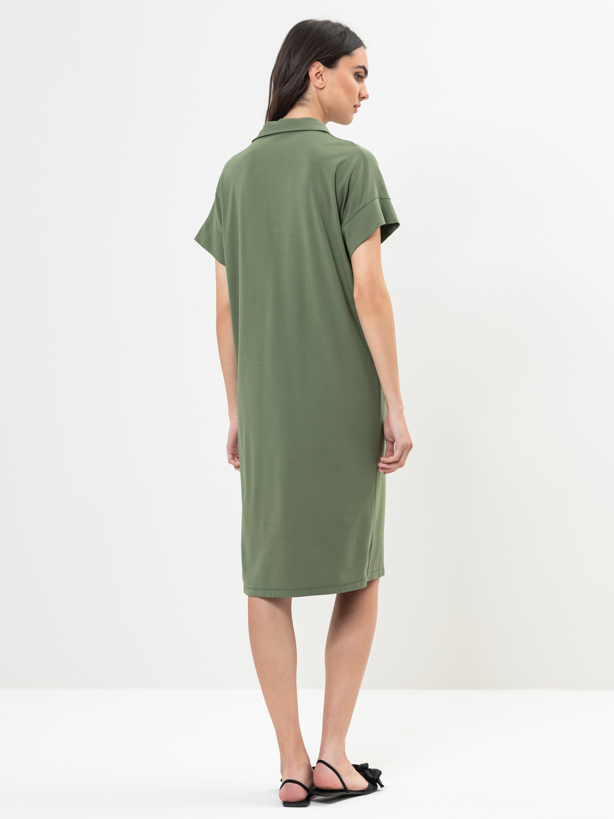 Eine Frau steht mit dem Gesicht zur Wand und trägt ein knielanges, kurzärmeliges olivgrünes Kleid mit einem Kragen. Sie hat langes dunkles Haar und trägt schwarze flache Sandalen. Der Hintergrund ist einfarbig weiß.