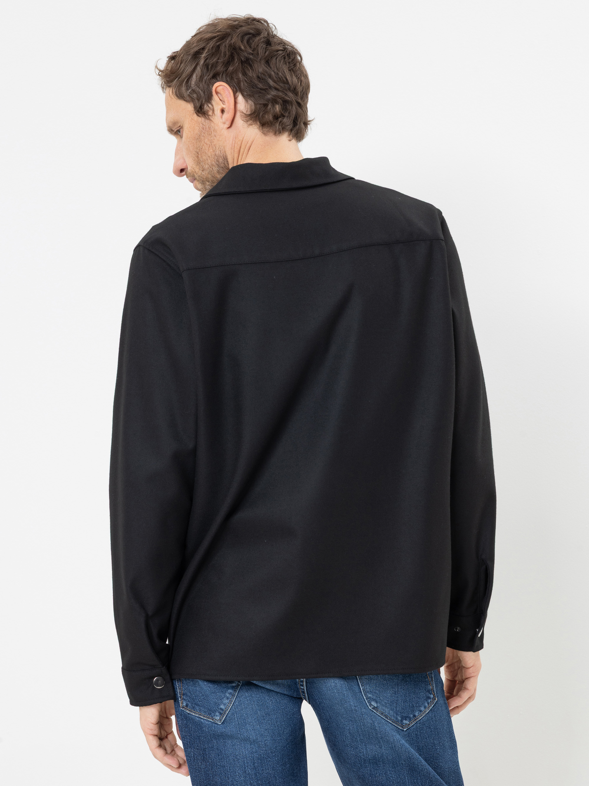 Vor einem schlichten weißen Hintergrund steht ein Mann mit kurzen braunen Haaren mit dem Rücken zur Kamera; er trägt ein schwarzes Langarmhemd und blaue Jeans.