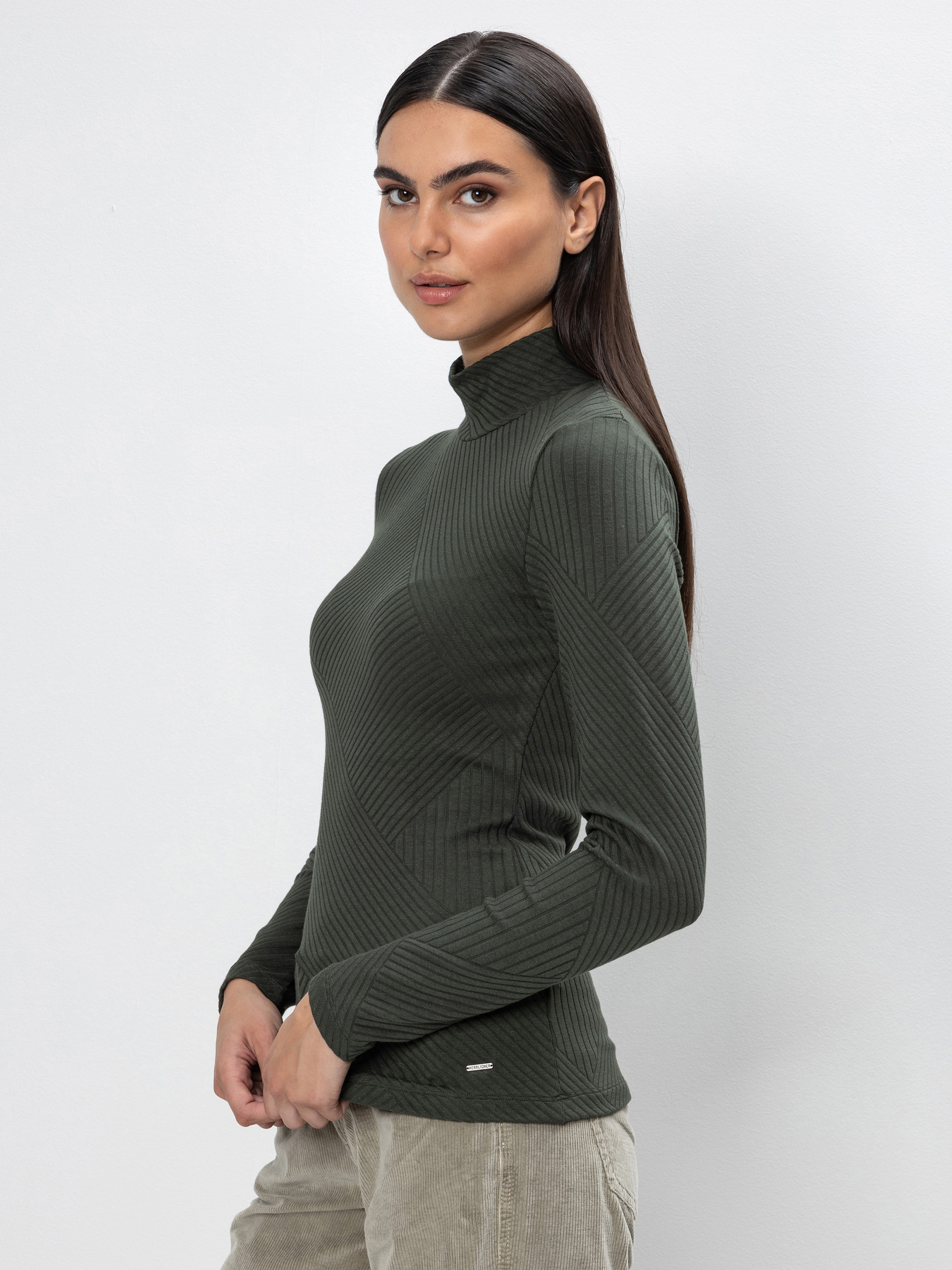 Eine Frau mit langem, glattem dunklem Haar posiert seitlich und trägt einen olivgrünen, gerippten, langärmeligen Rollkragenpullover und eine beigefarbene Hose vor einem schlichten hellgrauen Hintergrund.