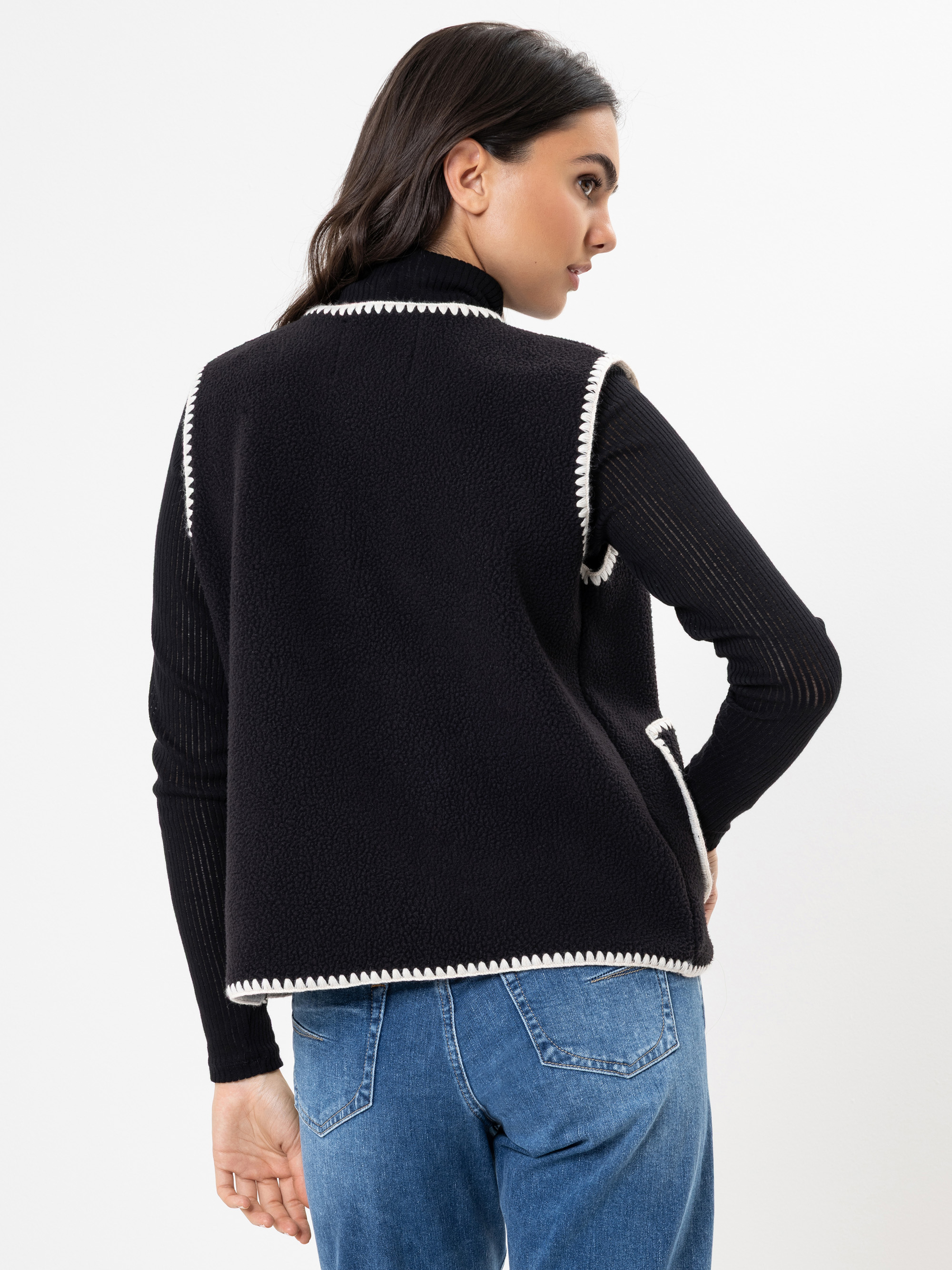 Eine Frau mit langen dunklen Haaren, bekleidet mit einem schwarzen langärmeligen Oberteil, einer schwarzen Weste mit weißem Saum und blauen Jeans, steht mit zur Seite gedrehtem Kopf vor einem weißen Hintergrund.