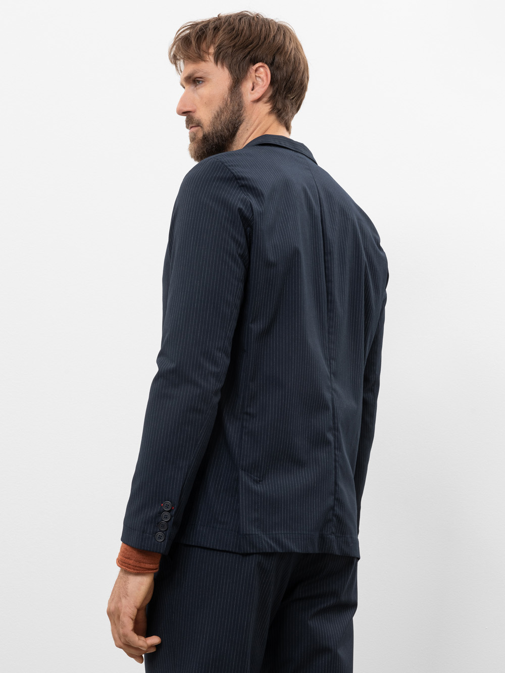 Ein Mann mit kurzen braunen Haaren und einem Bart trägt einen dunklen Nadelstreifenanzug und ein orange-braunes Hemd. Er steht seitlich und schaut über seine linke Schulter vor einem schlichten weißen Hintergrund.