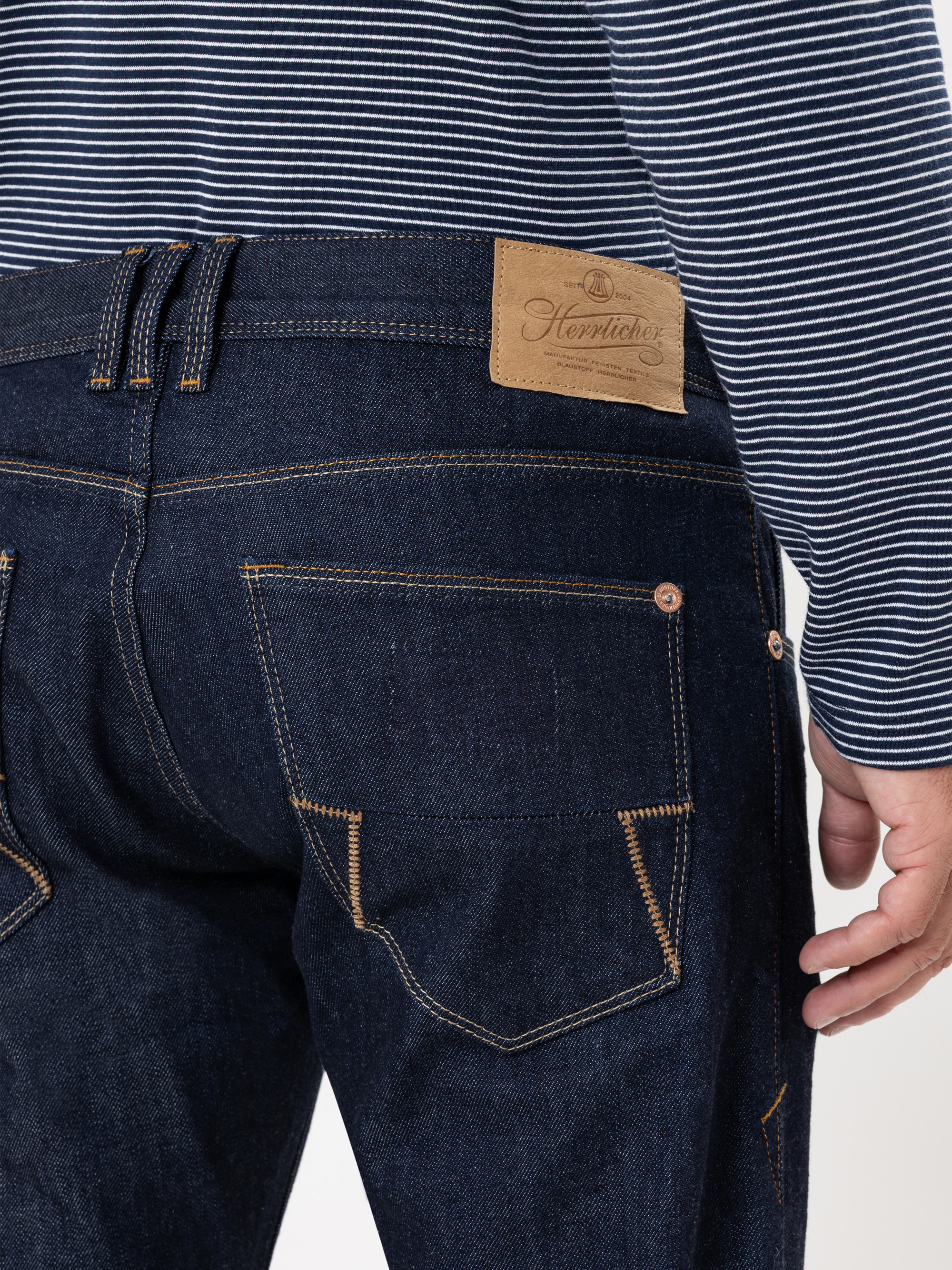 Eine Person trägt eine dunkelblaue Jeans und ein marineblau-weiß gestreiftes Hemd, von hinten gesehen. Die Jeans hat sichtbare braune Nähte, einen Markenaufnäher aus Leder und Gesäßtaschen mit markanten Nähten.