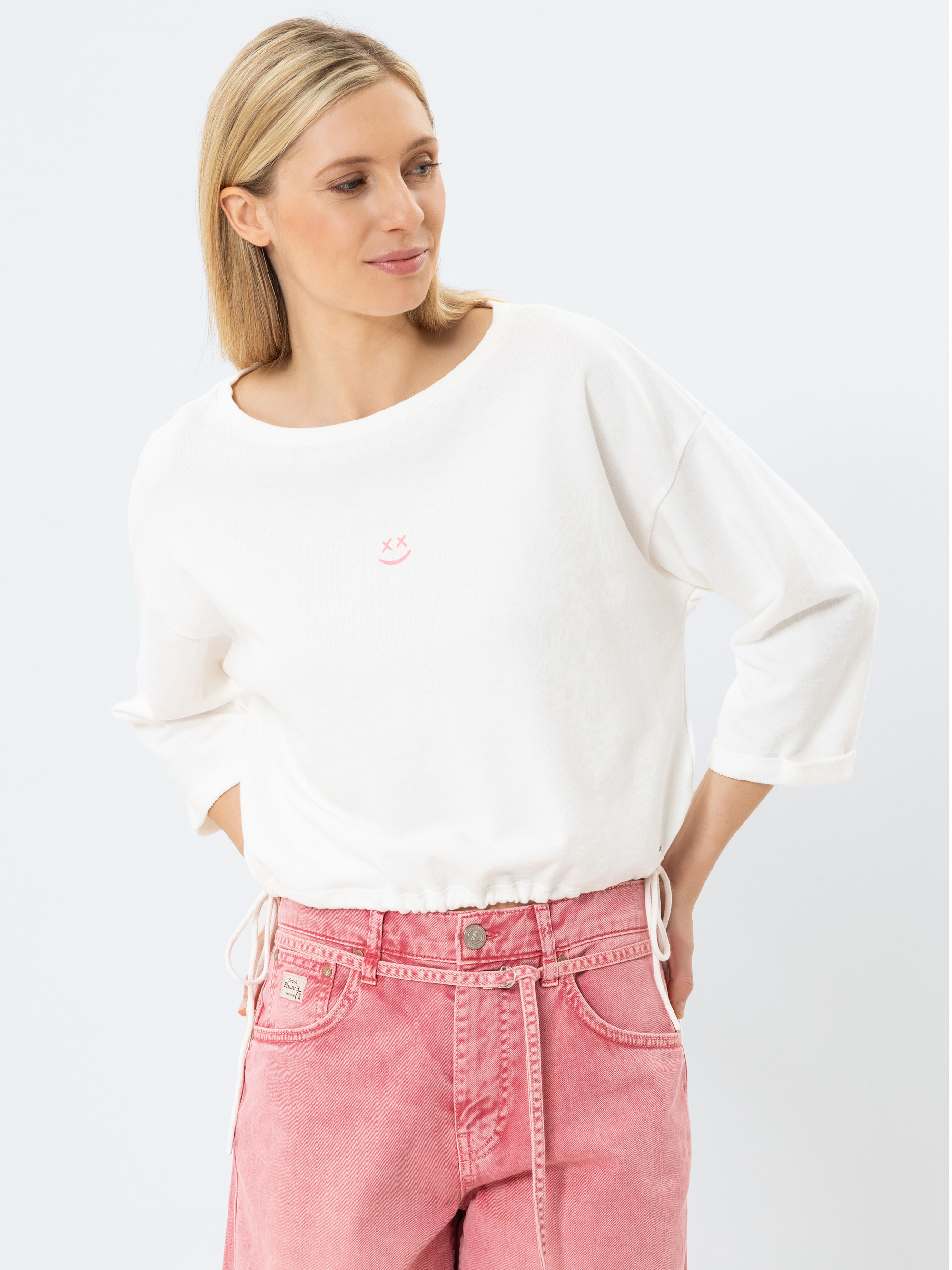 Eine Frau mit glattem blondem Haar trägt ein lockeres weißes langärmeliges Oberteil mit einer kleinen rosa Smiley-Grafik und hochgeschnittene rosa Jeans, die vor einem schlichten hellen Hintergrund stehen.