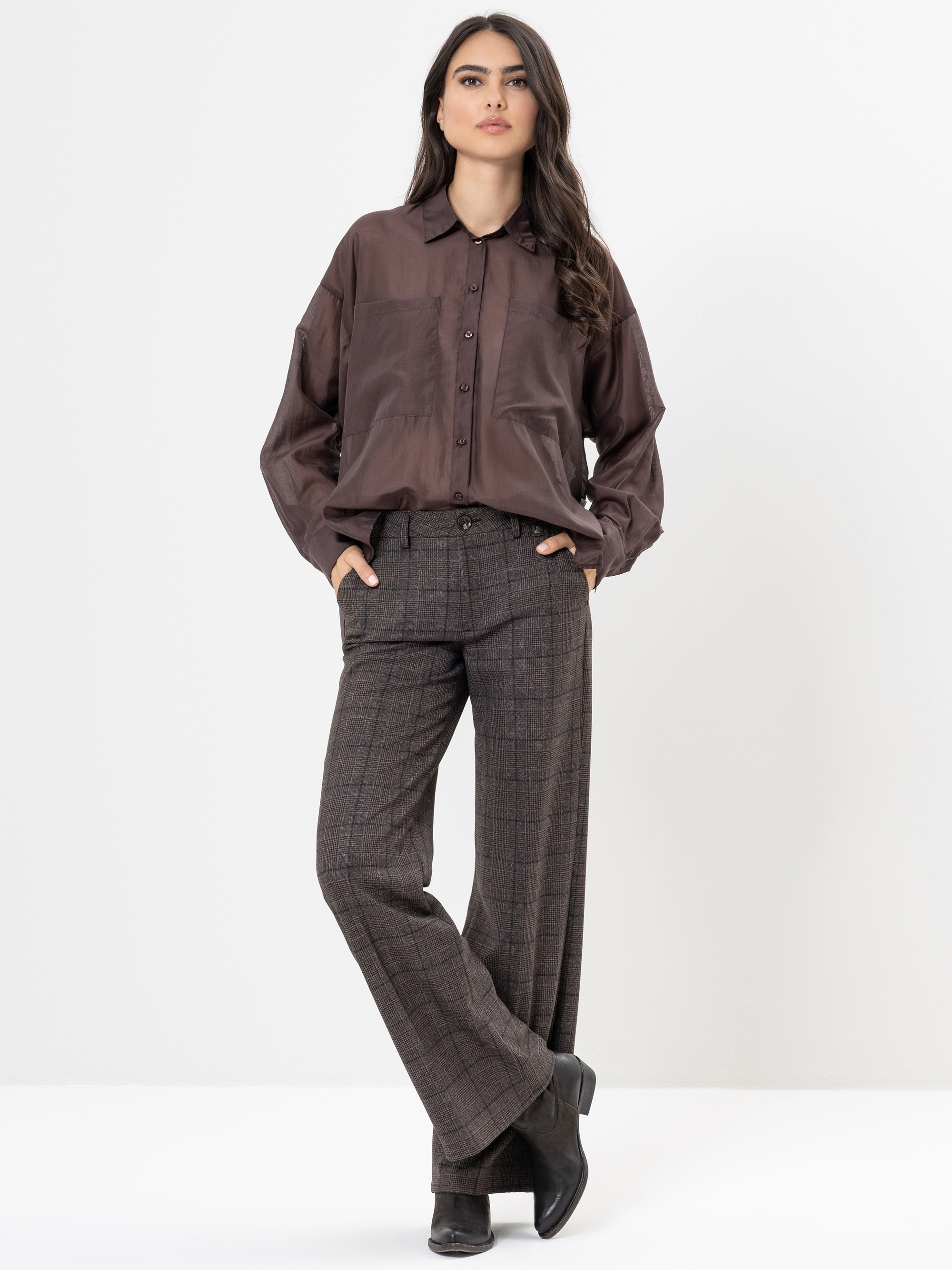 Eine Frau steht vor einem weißen Hintergrund und trägt ein lockeres, dunkelbraunes Button-up-Hemd und eine weite, hoch sitzende karierte Hose mit schwarzen Stiefeln. Sie hat langes, gewelltes Haar und ihre Hände stecken in den Taschen.