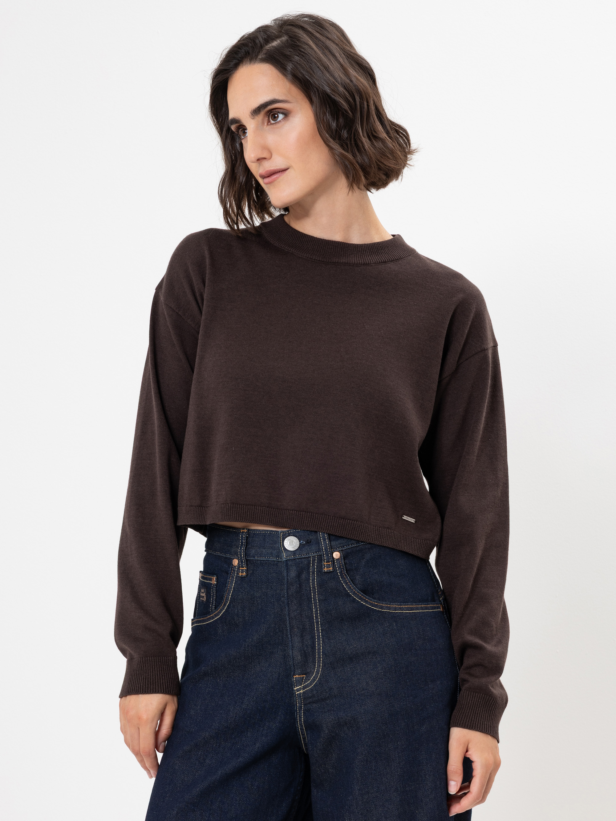 Eine Frau mit kurzen braunen Haaren, die einen dunkelbraunen, langärmeligen Pullover und hochgekrempelte dunkelblaue Jeans trägt, steht vor einem schlichten weißen Hintergrund.