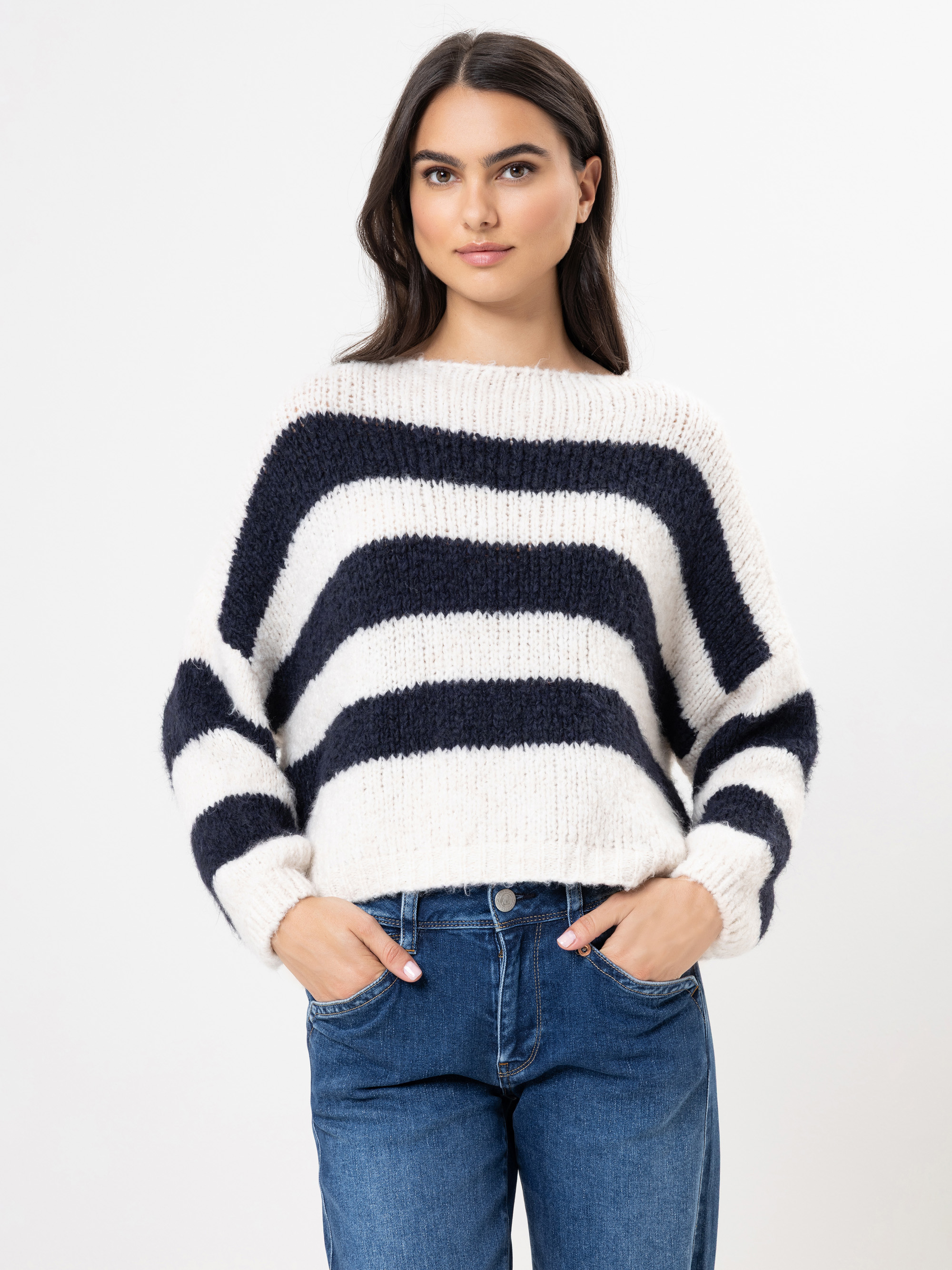 Eine Frau mit langen dunklen Haaren trägt einen wuscheligen weißen Pullover mit dicken schwarzen Querstreifen und blaue Jeans. Sie steht mit den Händen in den Taschen vor einem schlichten weißen Hintergrund.