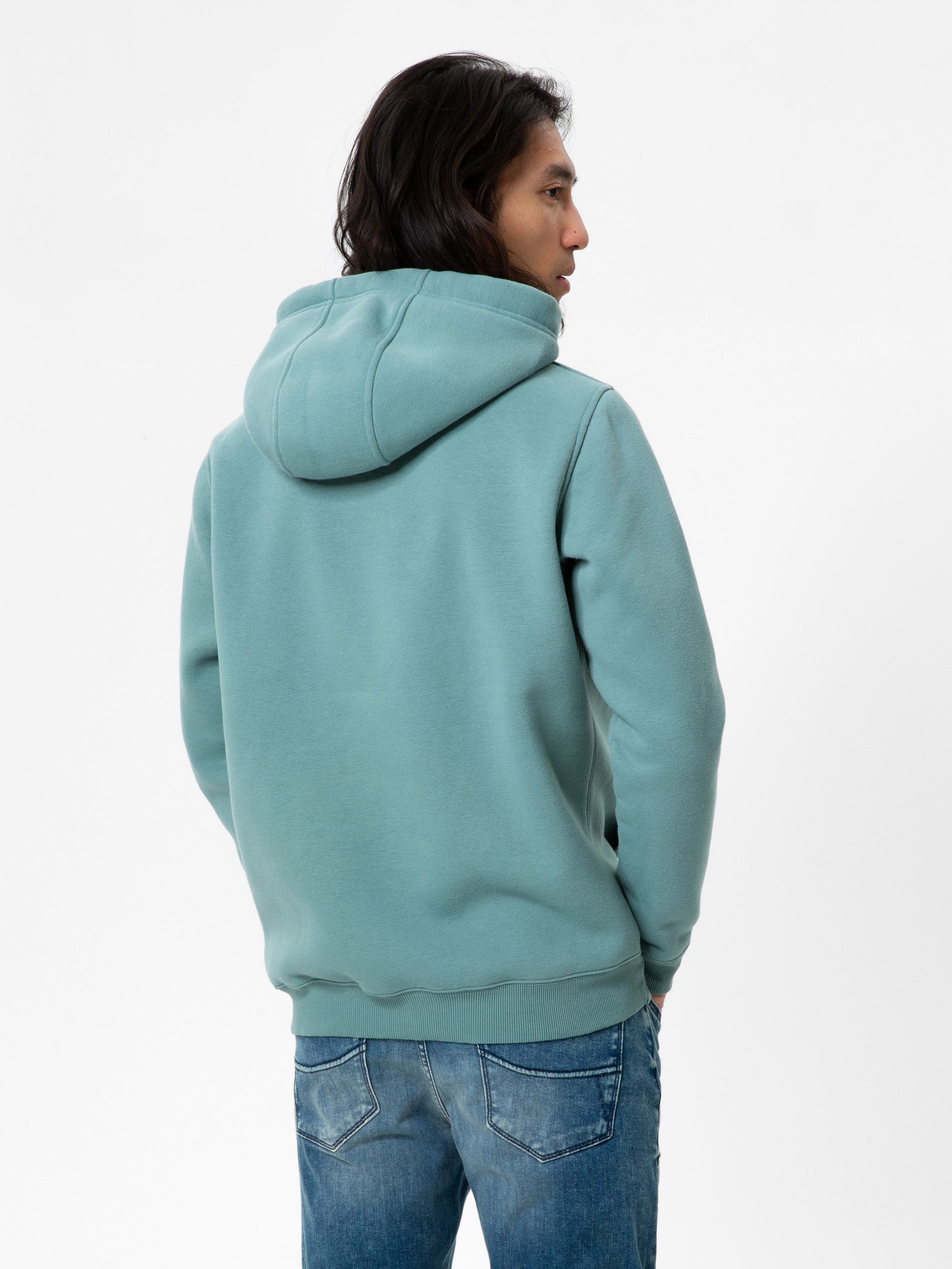 Eine Person mit langen dunklen Haaren, die einen hellgrünen Kapuzenpulli und blaue Jeans trägt, steht mit dem Gesicht zur Wand vor einem schlichten weißen Hintergrund.