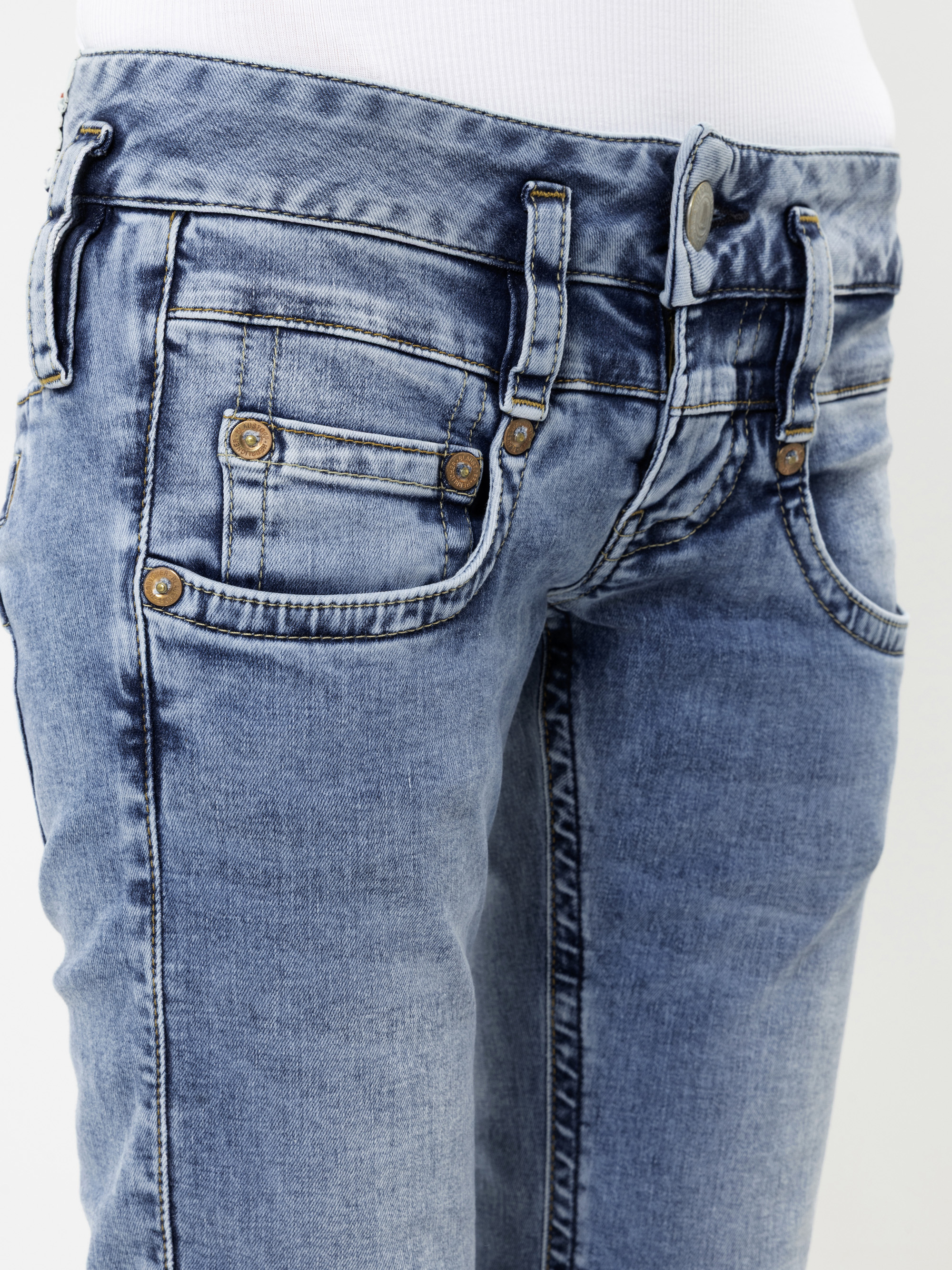 Nahaufnahme einer Person, die hellblaue Jeans und ein weißes Oberteil trägt. Die Jeans weist sichtbare Nähte, einen Knopfverschluss, Vordertaschen, Kupfernieten sowie eine verwaschene, leicht abgenutzte Optik auf.