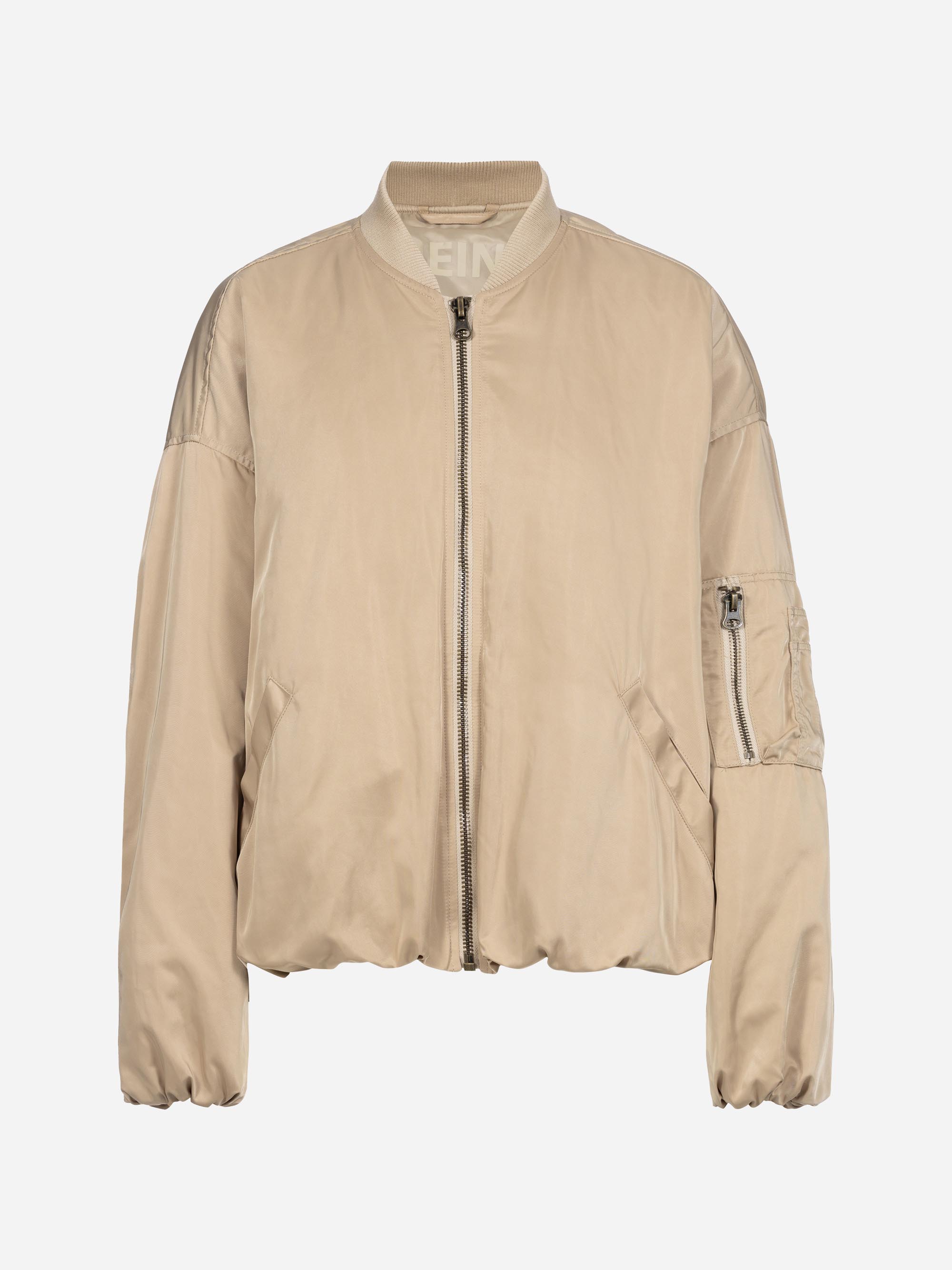 Beigefarbene Bomberjacke mit Frontreißverschluss, geripptem Kragen, Manschetten und Saum, zwei Seitentaschen und einer Reißverschlusstasche am linken Ärmel. Der Stoff wirkt glatt und leicht glänzend.