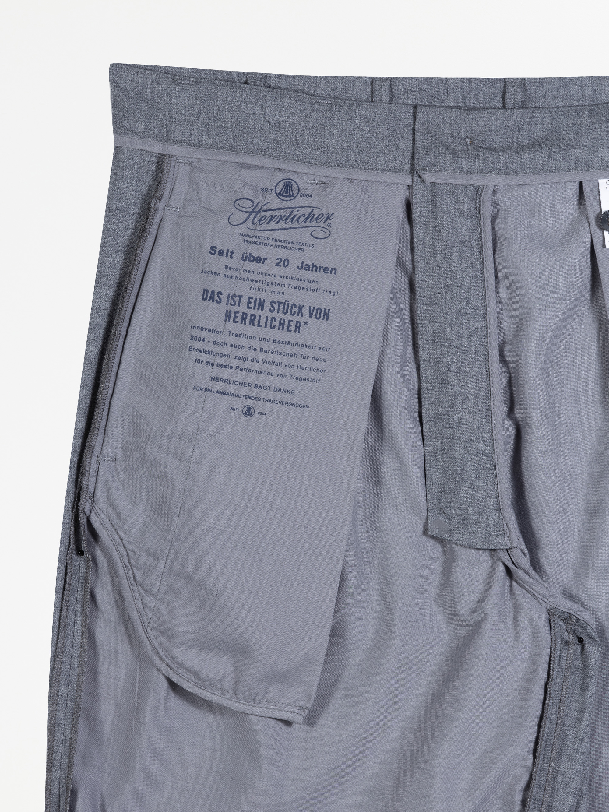 Nahaufnahme der Innenseite einer grauen Hose mit detaillierten Nähten, Taschenfutter und gedrucktem Text in deutscher Sprache, der die Marke Herrlicher und Pflegehinweise angibt.