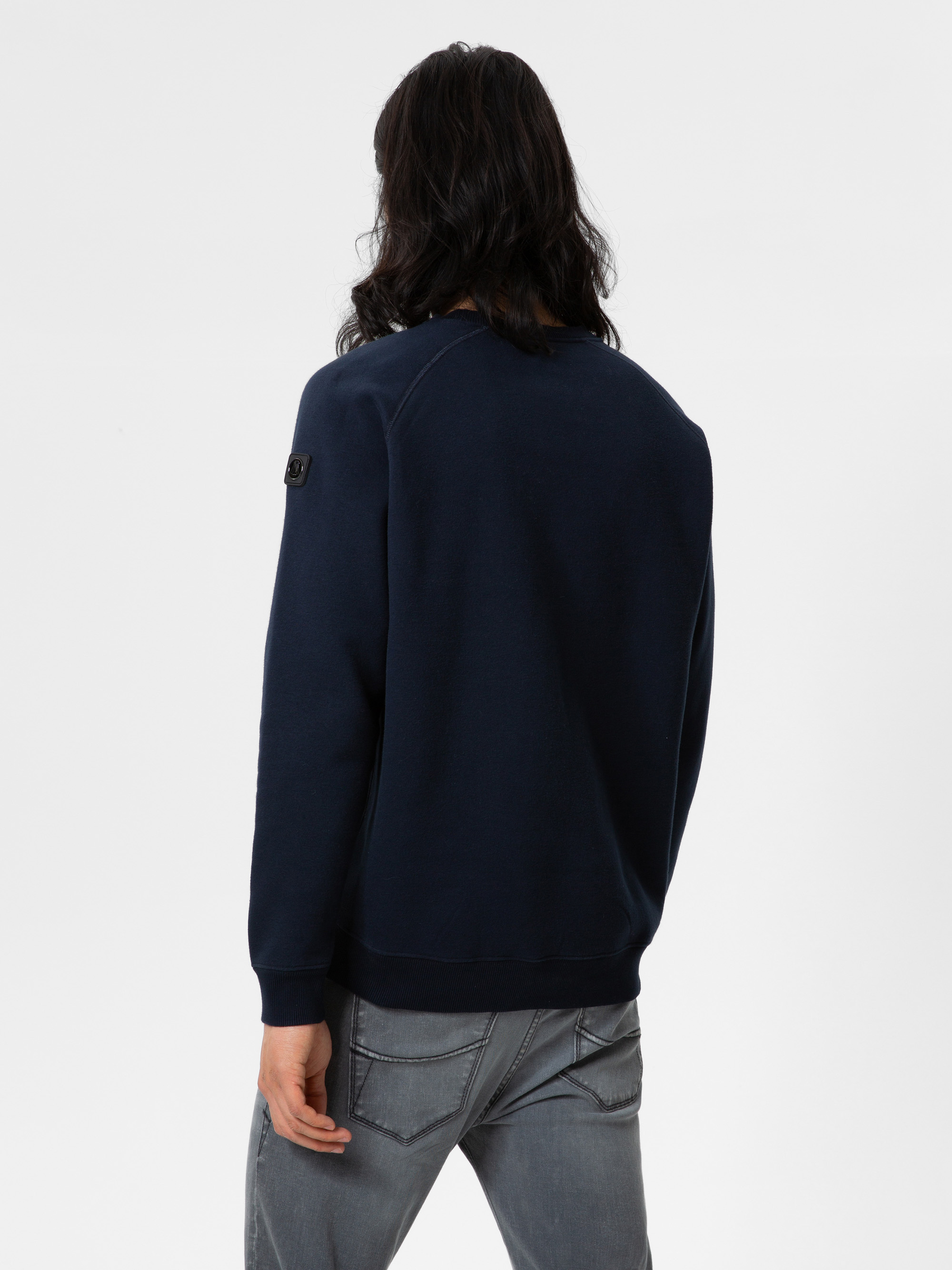 Eine Person mit langen dunklen Haaren, die ein marineblaues Sweatshirt und graue Jeans trägt, steht mit dem Gesicht zur Kamera vor einem schlichten weißen Hintergrund.