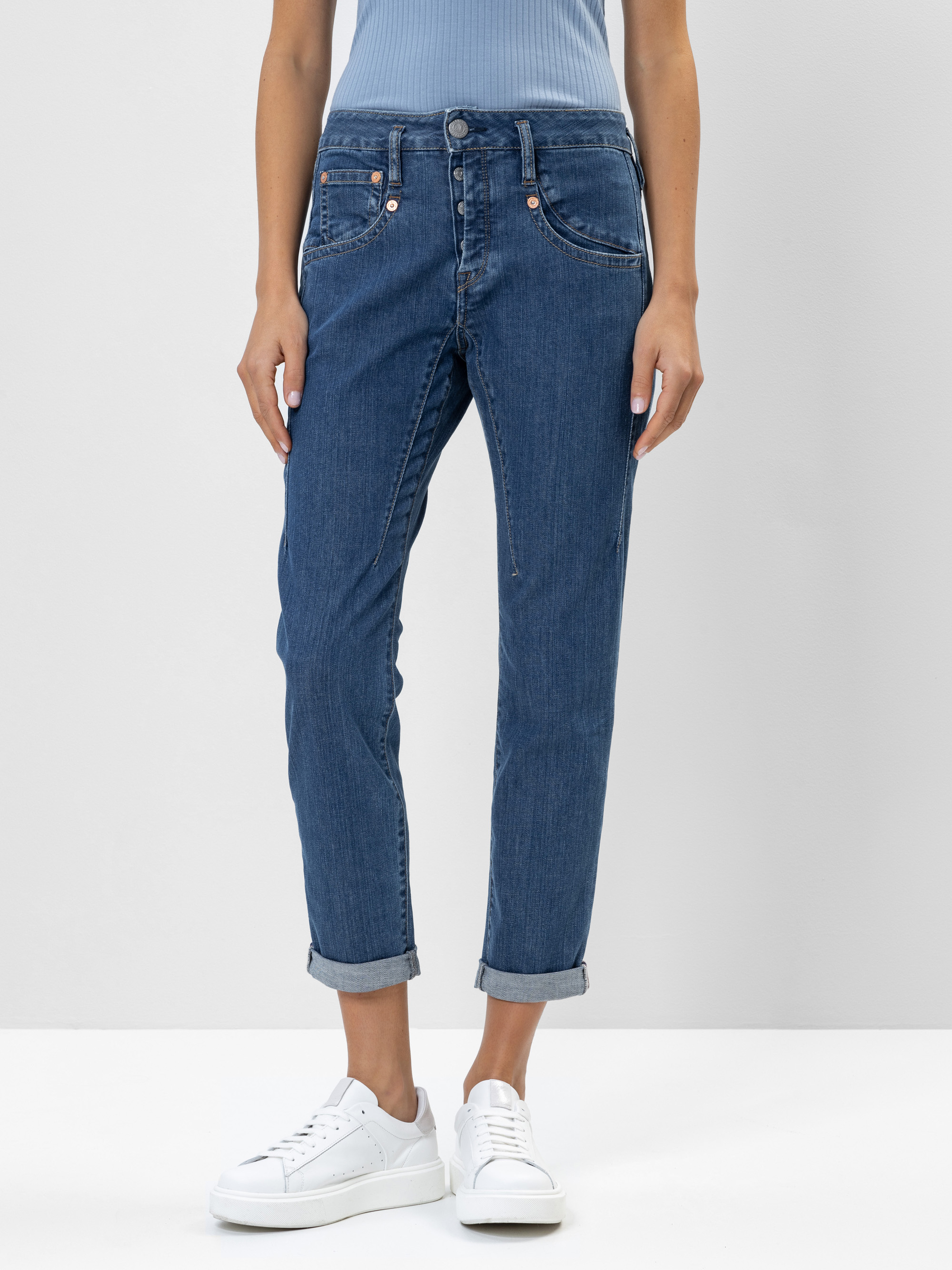 Eine Person, die eine blaue Jeans mit Bündchen, ein hellblaues geripptes Oberteil und weiße Turnschuhe trägt, steht vor einem schlichten hellen Hintergrund. Das Bild ist von den Schultern abwärts beschnitten.