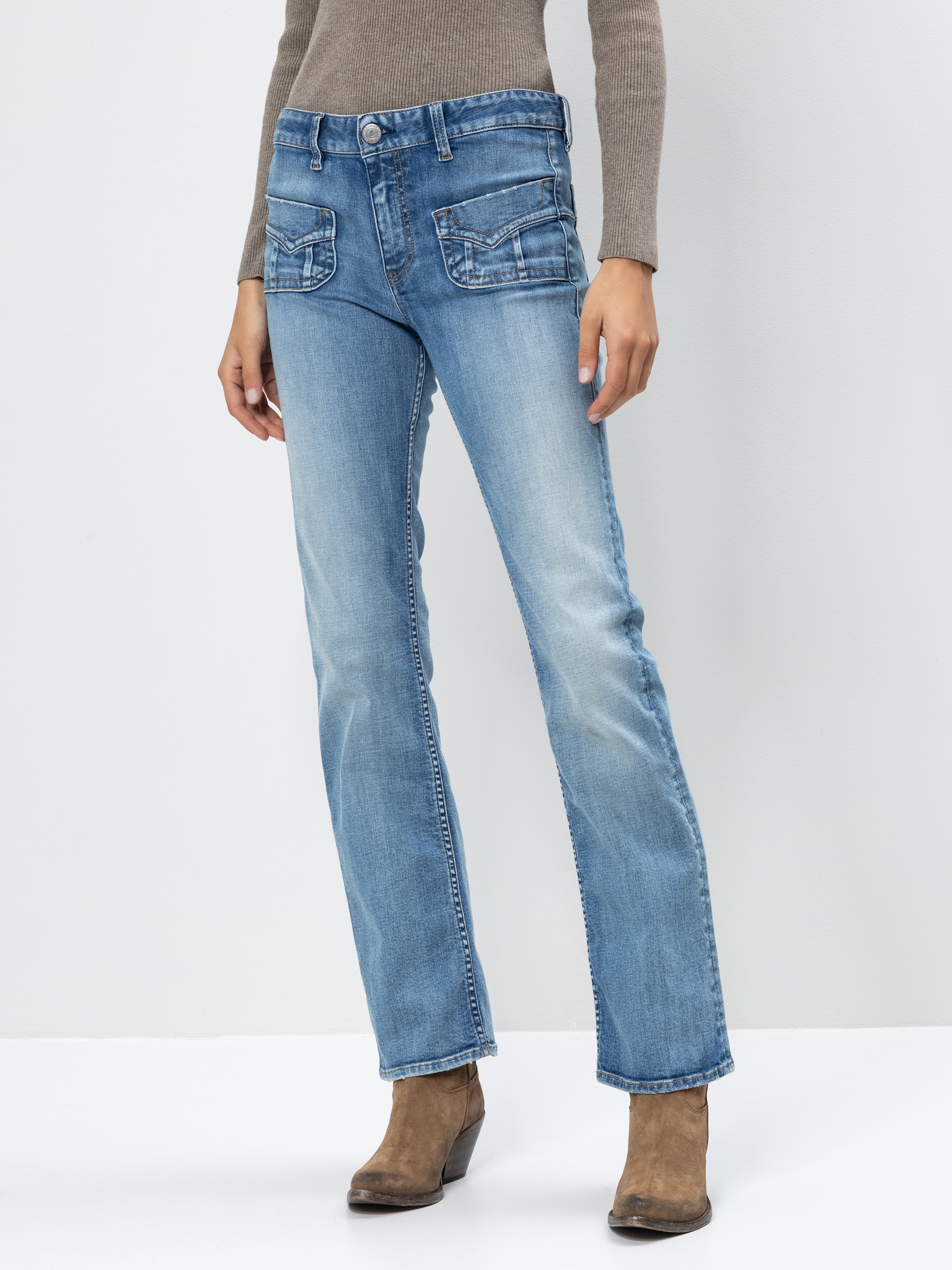 Eine Frau trägt hellblaue Jeans mit geradem Bein und Klappentaschen, einen taupefarbenen gerippten Pullover und braune Wildlederstiefel vor einem schlichten weißen Hintergrund.