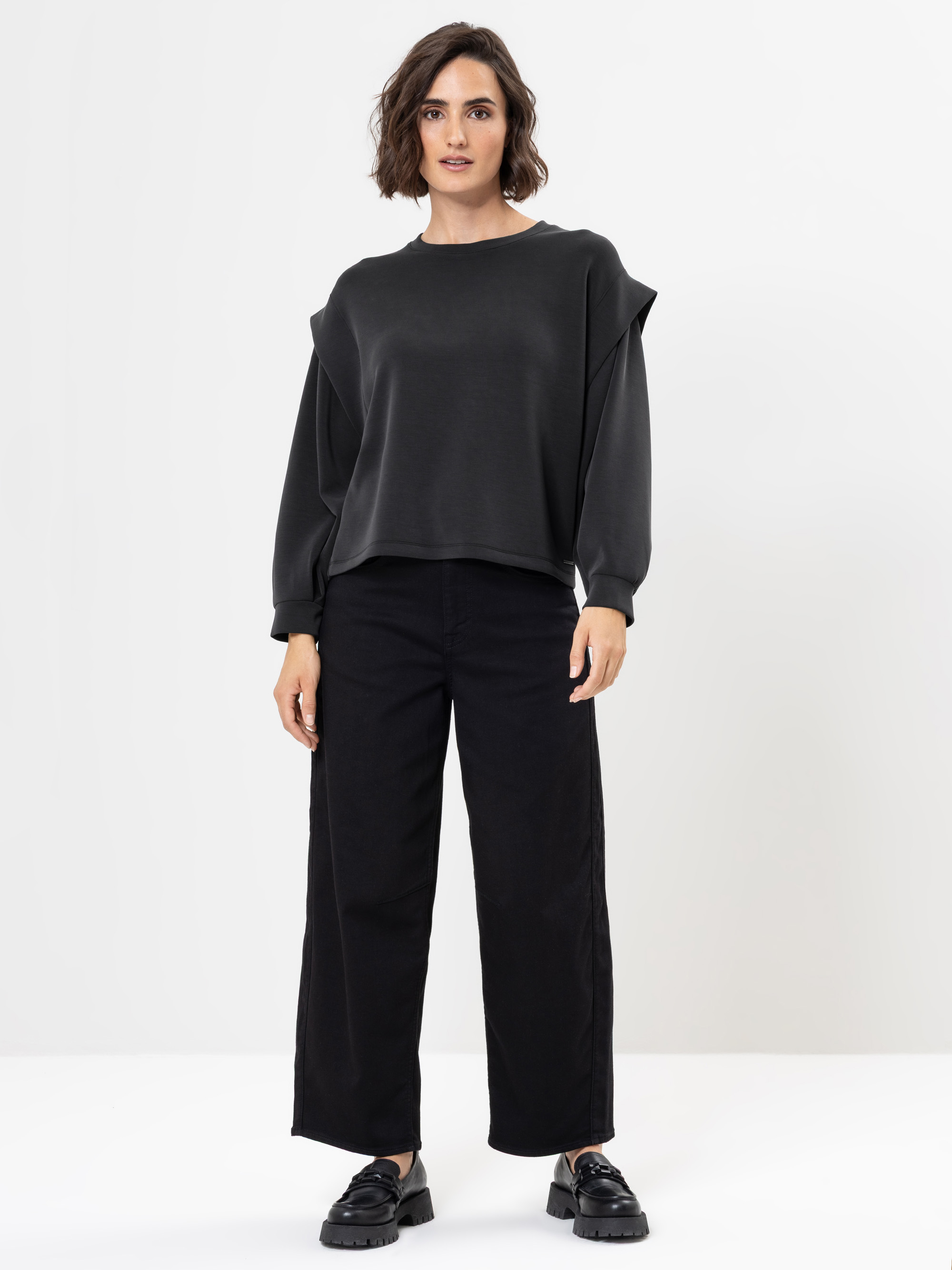 Eine Frau mit kurzen braunen Haaren trägt einen weit geschnittenen schwarzen Pullover, eine schwarze Hose mit weitem Bein und schwarze Schuhe und steht vor einem schlichten weißen Hintergrund.