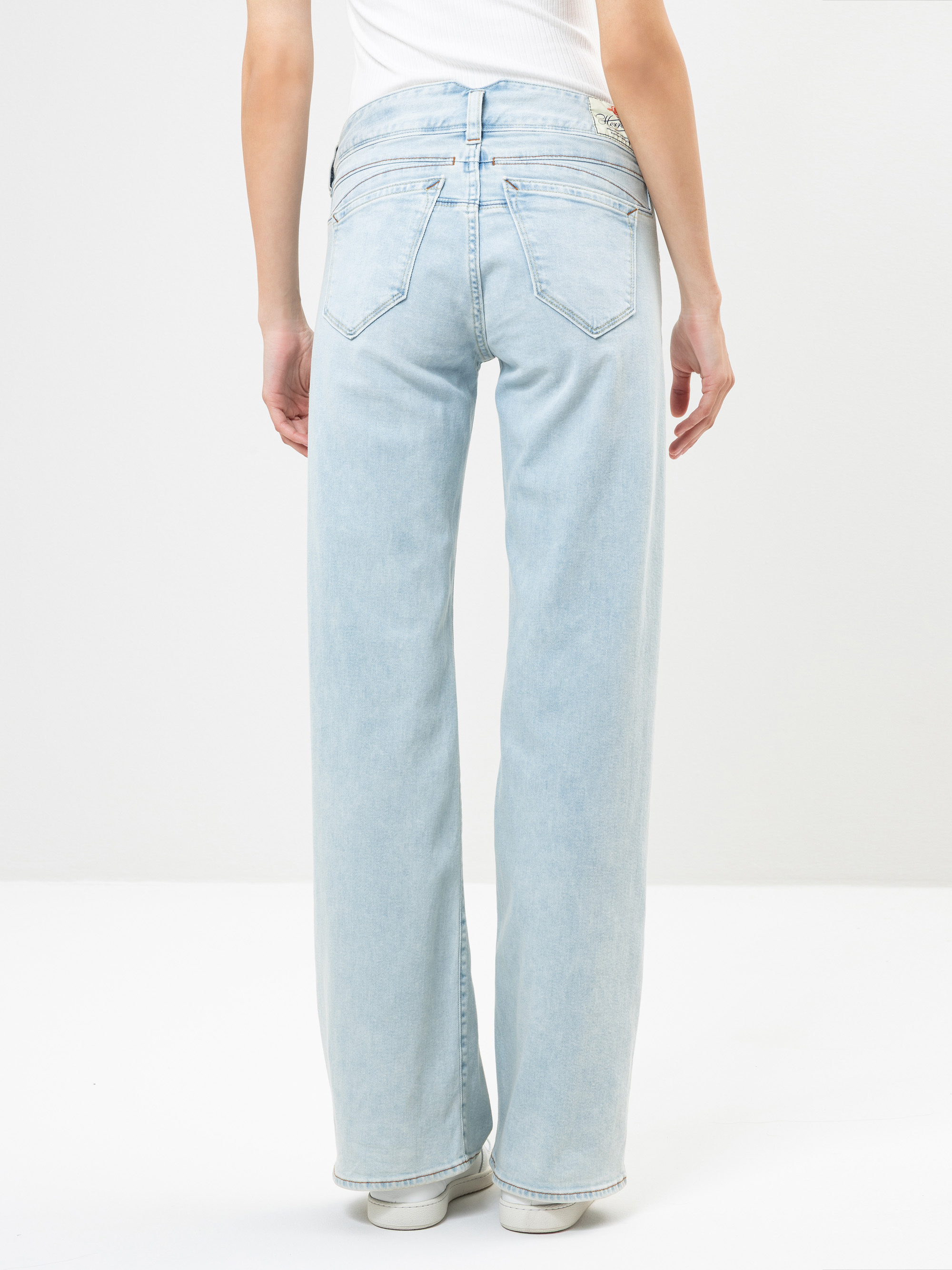 Eine Person, die hellblaue Jeans mit weitem Bein und ein weißes ärmelloses Oberteil trägt, steht mit dem Rücken zur Kamera vor einem schlichten weißen Hintergrund.