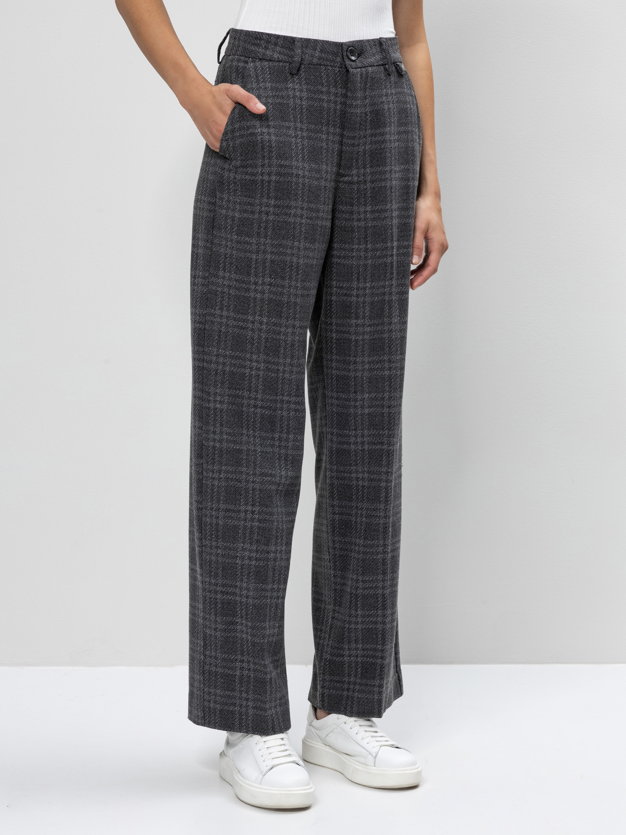 Eine Person, die eine grau karierte Hose mit weitem Bein, ein weißes ärmelloses Oberteil und weiße Turnschuhe trägt, steht mit einer Hand in der Tasche vor einem schlichten hellen Hintergrund.