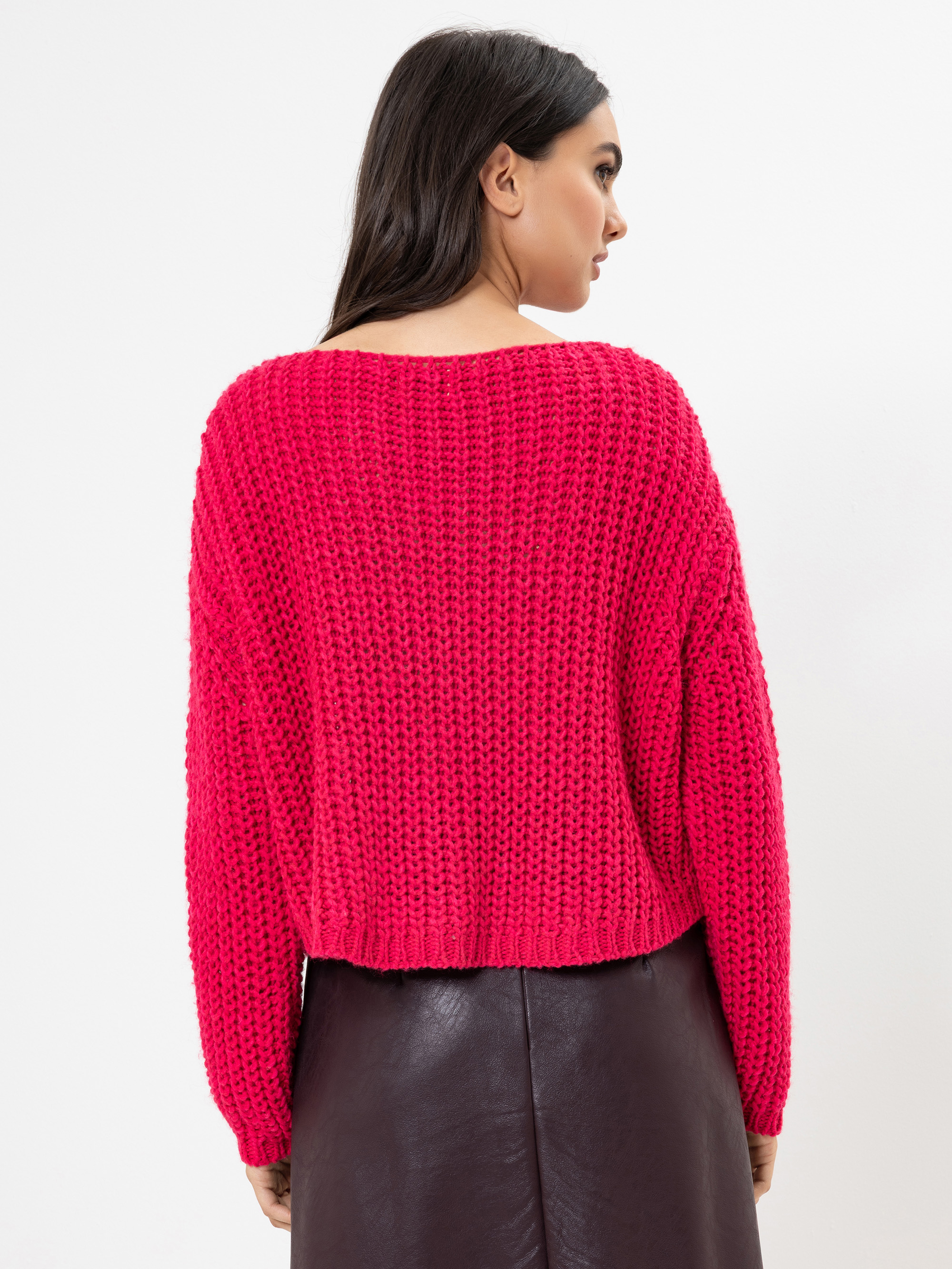 Eine Frau steht mit dem Rücken zur Kamera und trägt einen hellrosa Strickpullover und einen dunklen Lederrock vor einem schlichten weißen Hintergrund.