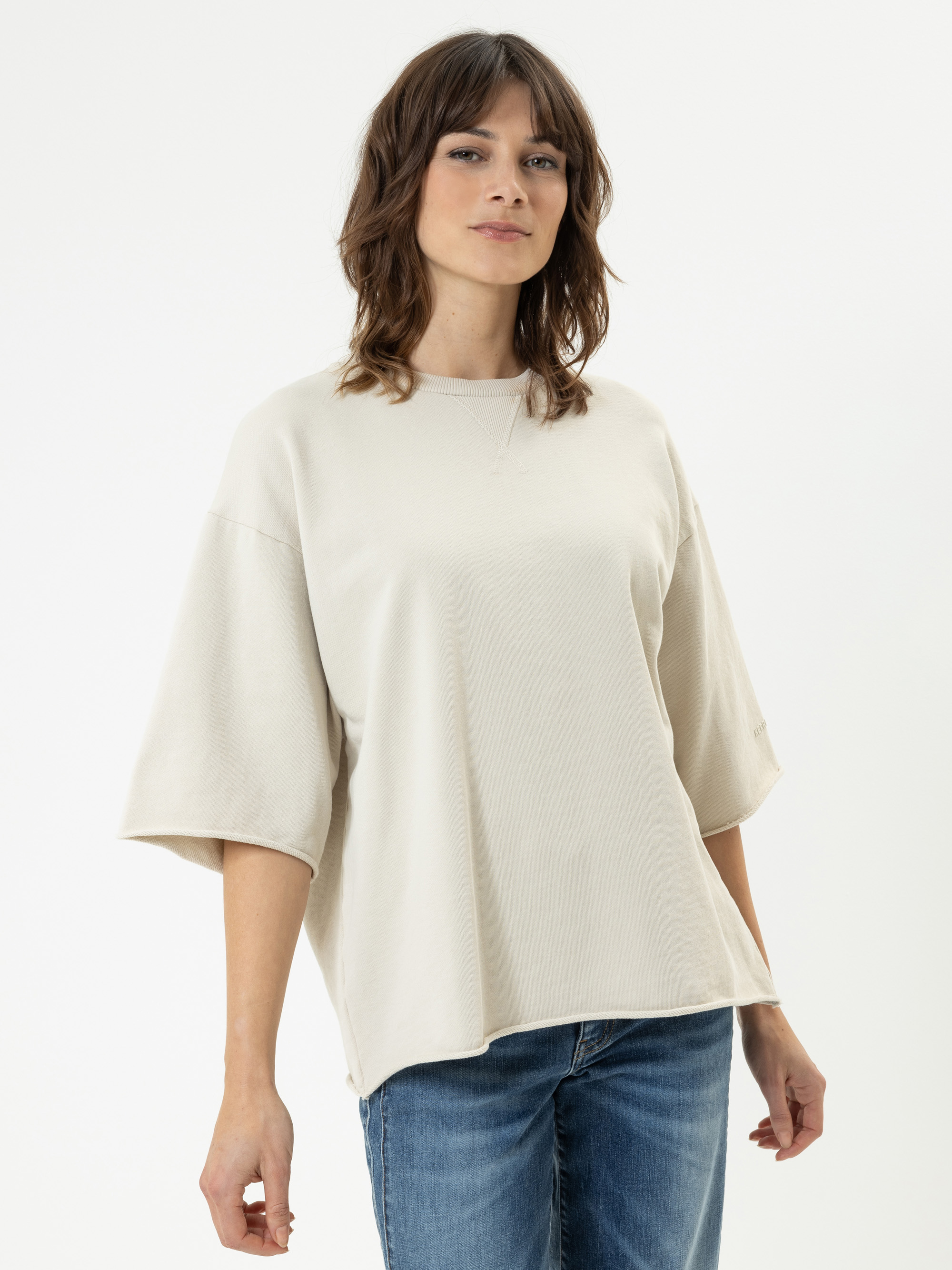 Eine Frau mit mittellangem braunem Haar, die ein lockeres, hellbeiges, kurzärmeliges Oberteil und blaue Jeans trägt, steht vor einem schlichten weißen Hintergrund und blickt selbstbewusst in die Kamera.