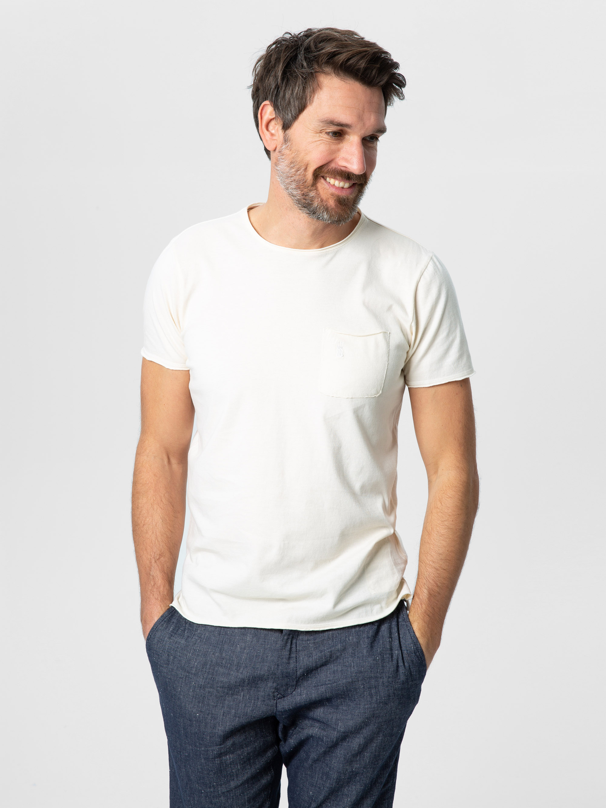 Ein Mann mit kurzen braunen Haaren und Bart, bekleidet mit einem weißen kurzärmeligen T-Shirt und einer blauen Hose, steht mit den Händen in den Taschen, lächelt und schaut leicht zur Seite vor einem schlichten hellen Hintergrund.