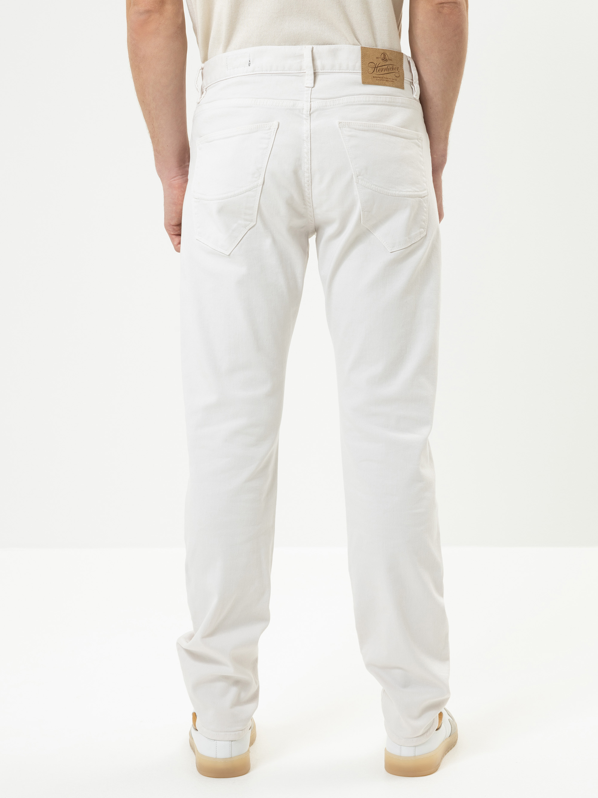 Eine Person steht mit dem Gesicht nach hinten, trägt weiße Jeans, ein beiges Oberteil und weiße Turnschuhe mit hellbraunen Sohlen vor einem schlichten weißen Hintergrund.