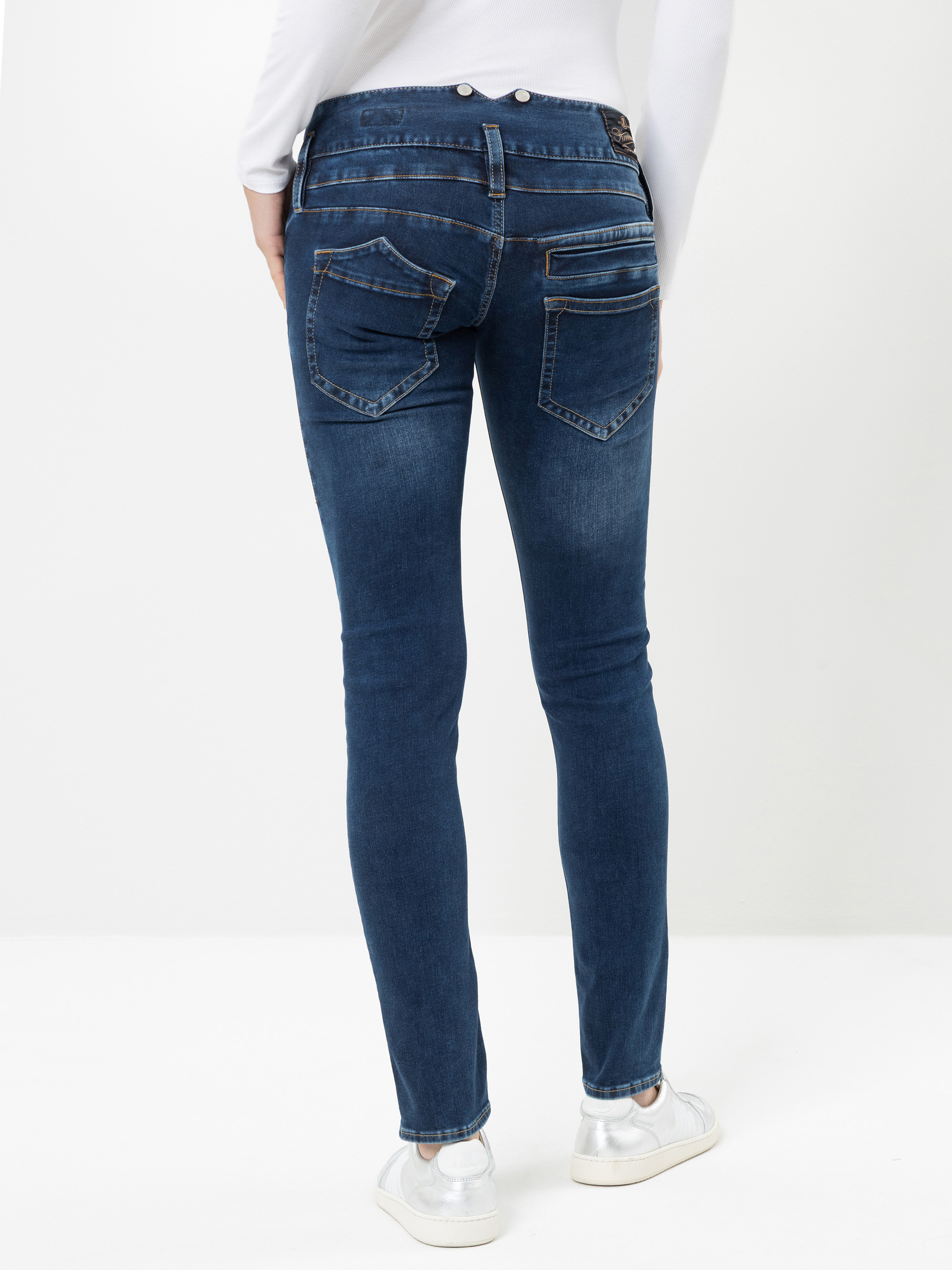 Eine Person, die ein weißes Oberteil und eine blaue Skinny-Jeans trägt, ist von hinten zu sehen. Die Jeans hat sichtbare Gesäßtaschen mit Ziernähten. Die Person trägt außerdem weiße Schuhe und steht auf einer weißen Fläche vor einem einfarbigen