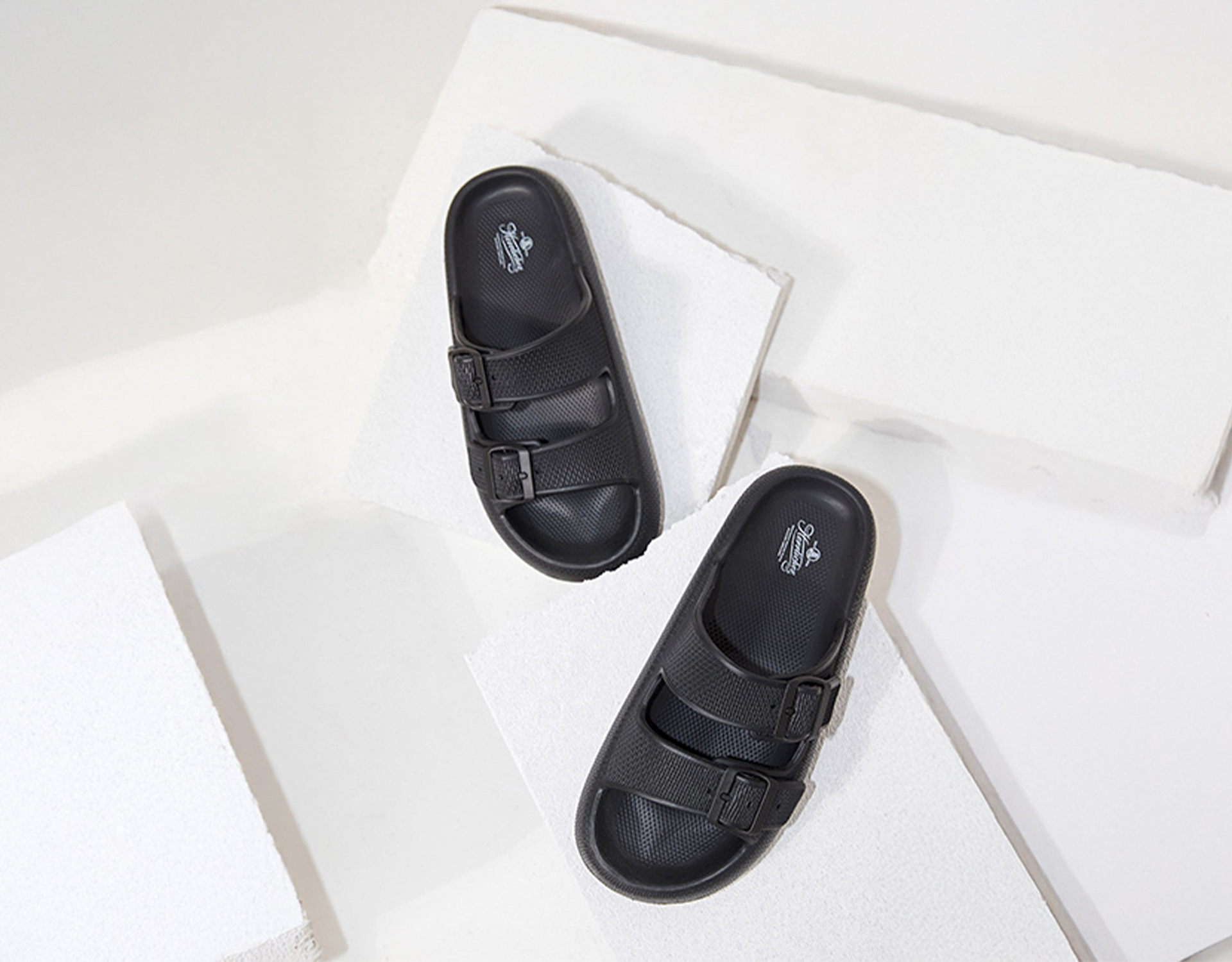 Ein Paar schwarze Schlupfsandalen mit zwei Schnallenriemen werden auf weißen, strukturierten Plateaus vor weißem Hintergrund präsentiert.