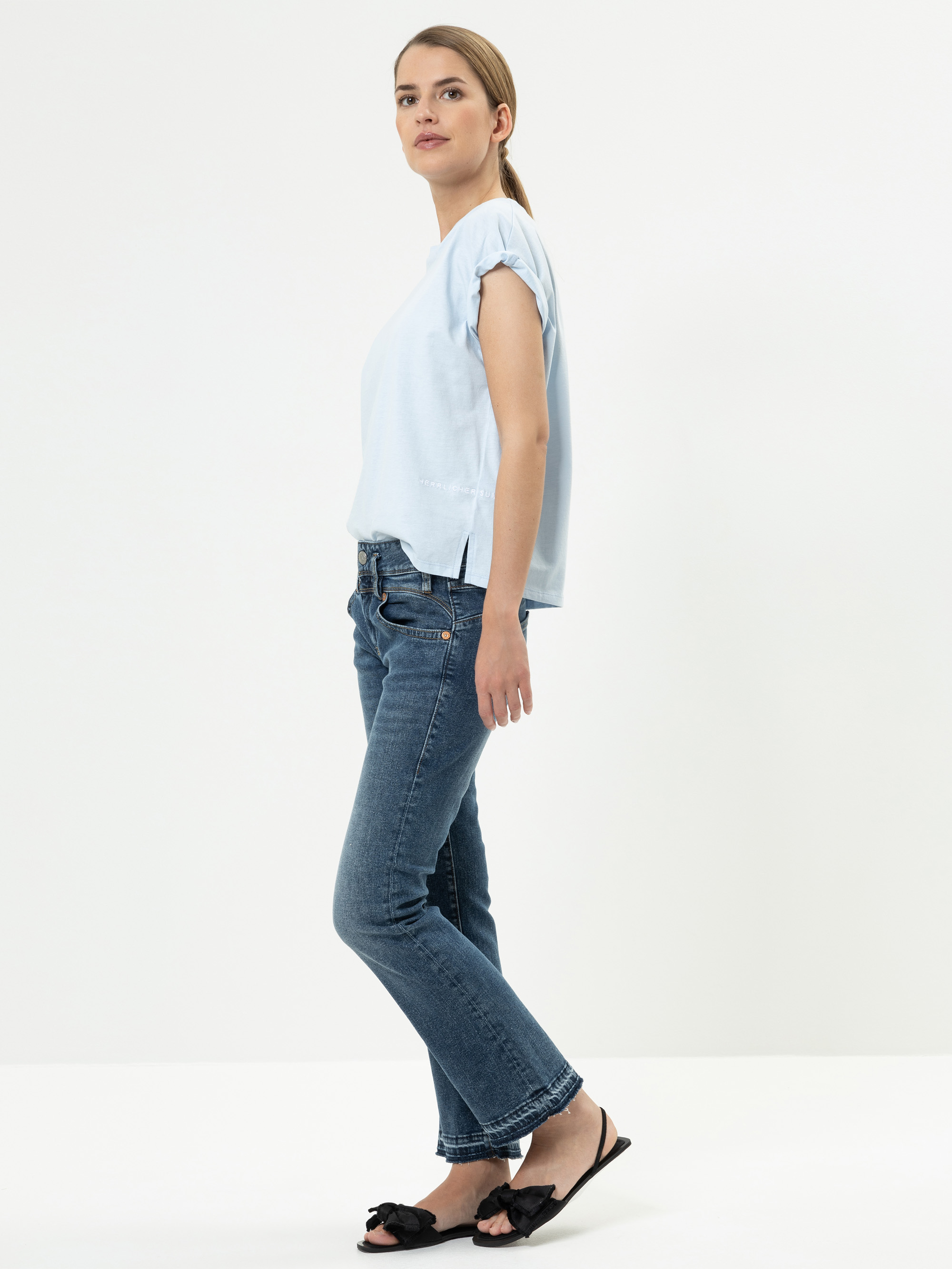 Eine Frau steht seitlich vor einem schlichten weißen Hintergrund. Sie trägt ein hellblaues kurzärmeliges Oberteil, blaue Jeans und schwarze Sandalen. Sie hat ihr hellbraunes Haar zurückgebunden.