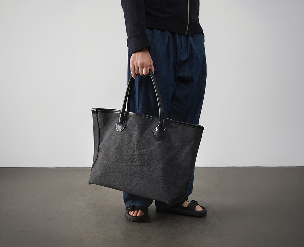 Eine Person in weiten blauen Hosen und schwarzen Sandalen hält eine große schwarze Tragetasche mit zwei Henkeln vor einem unifarbenen Hintergrund. Der Oberkörper der Person ist teilweise sichtbar.