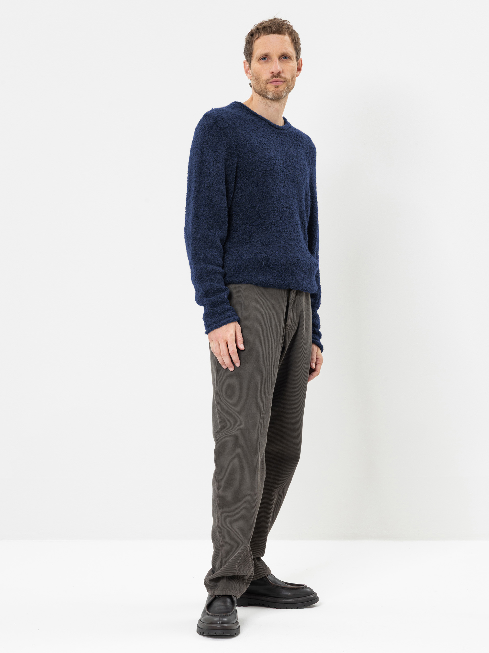 Ein Mann steht vor einem weißen Hintergrund und trägt einen strukturierten marineblauen Pullover, eine graue Hose und schwarze Schuhe. Er hat kurzes braunes Haar, einen leichten Bartwuchs und blickt mit neutralem Gesichtsausdruck leicht zur Seite.