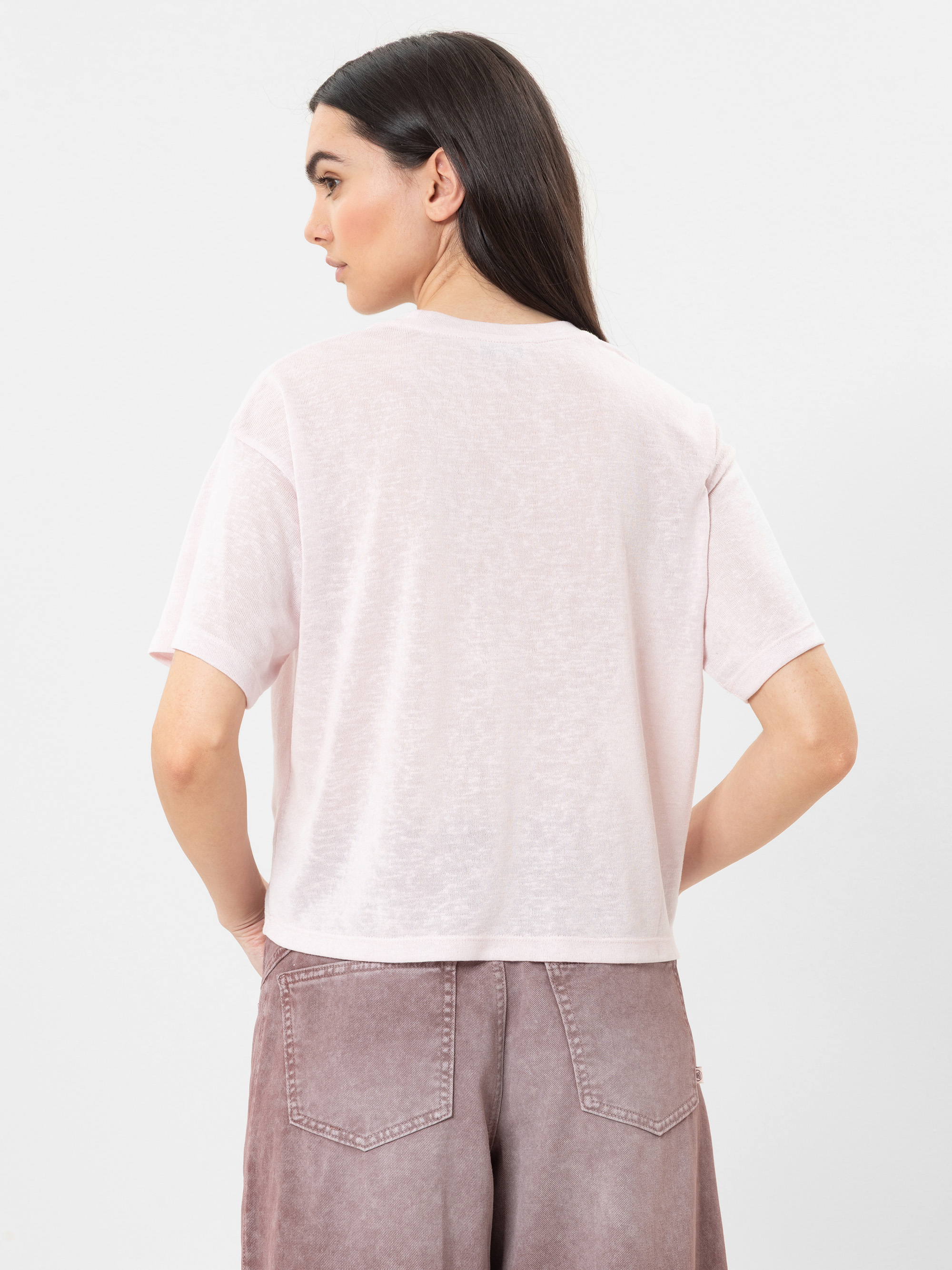 Eine Frau mit langen dunklen Haaren steht mit dem Rücken zur Kamera und trägt ein hellrosa, kurzärmeliges T-Shirt und lilafarbene, hochgekrempelte Jeans. Der Hintergrund ist einfarbig weiß.