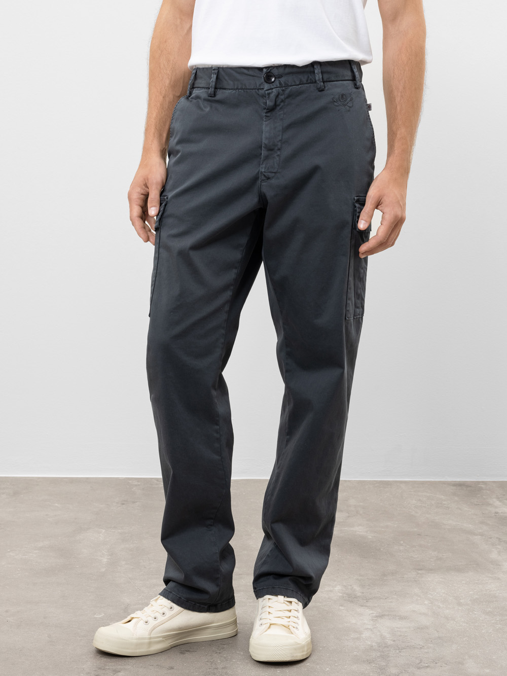 Eine Person, die ein weißes T-Shirt, eine dunkelgraue Cargohose und weiße Turnschuhe trägt, steht auf einem grauen Boden vor einem hellgrauen Hintergrund. Nur die untere Hälfte des Körpers ist sichtbar.