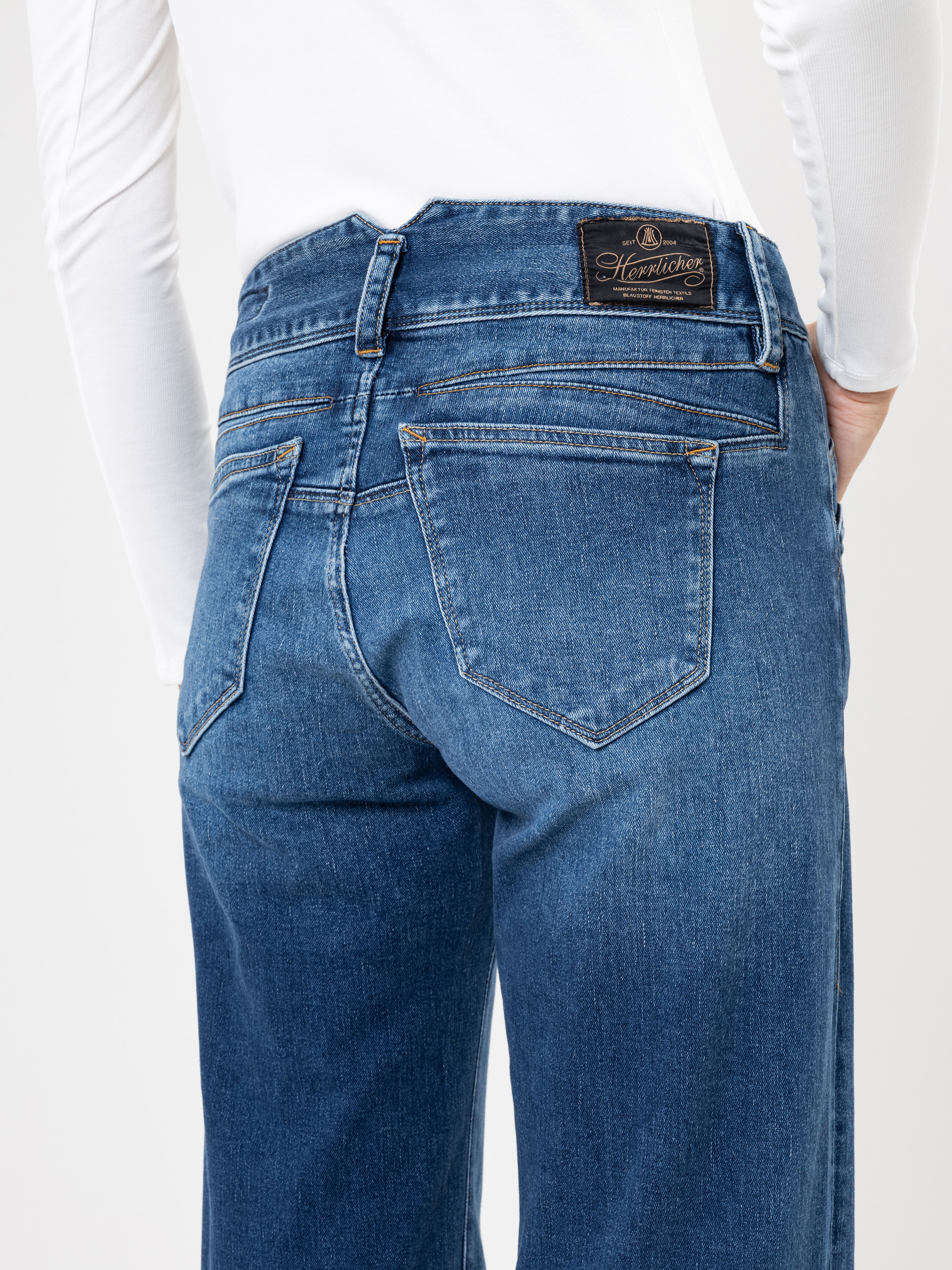Person, die eine blaue Jeans und ein langärmeliges weißes Oberteil trägt, dargestellt von der Taille abwärts und von hinten. Die Jeans hat einen braunen Lederaufnäher mit Branding am Bund über der rechten Gesäßtasche.