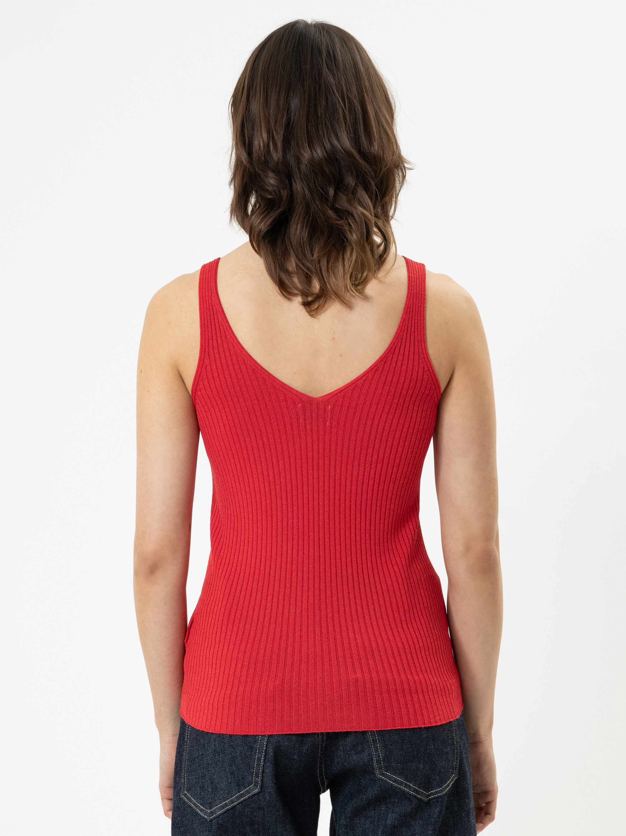 Eine Person mit schulterlangen braunen Haaren ist von hinten zu sehen. Sie trägt ein rotes, geripptes, ärmelloses Oberteil und dunkelblaue Jeans vor einem schlichten weißen Hintergrund.
