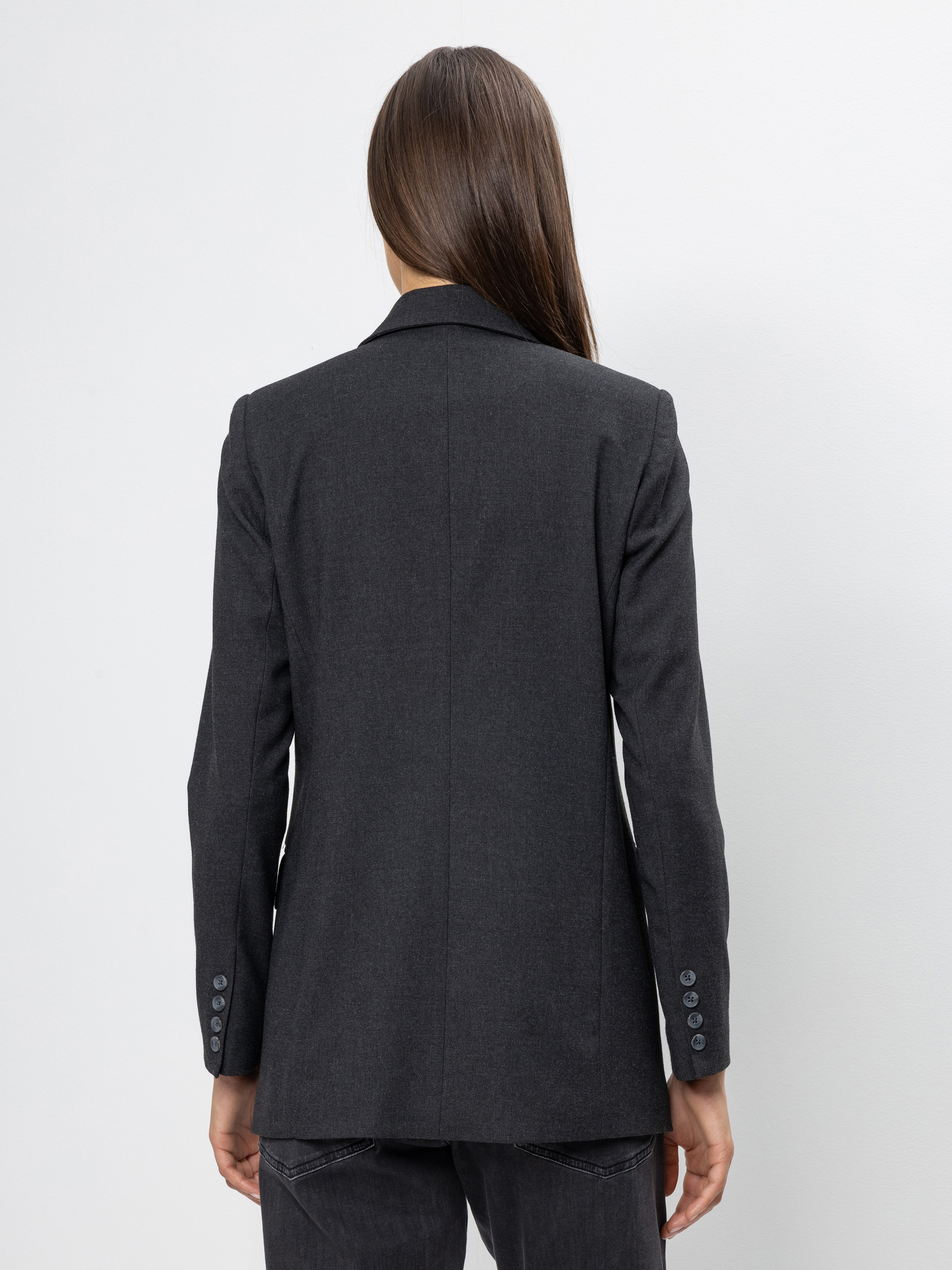 Eine Person mit langen braunen Haaren wird von hinten gezeigt, die einen dunkelgrauen Blazer und eine dazu passende Hose vor einem schlichten weißen Hintergrund trägt.