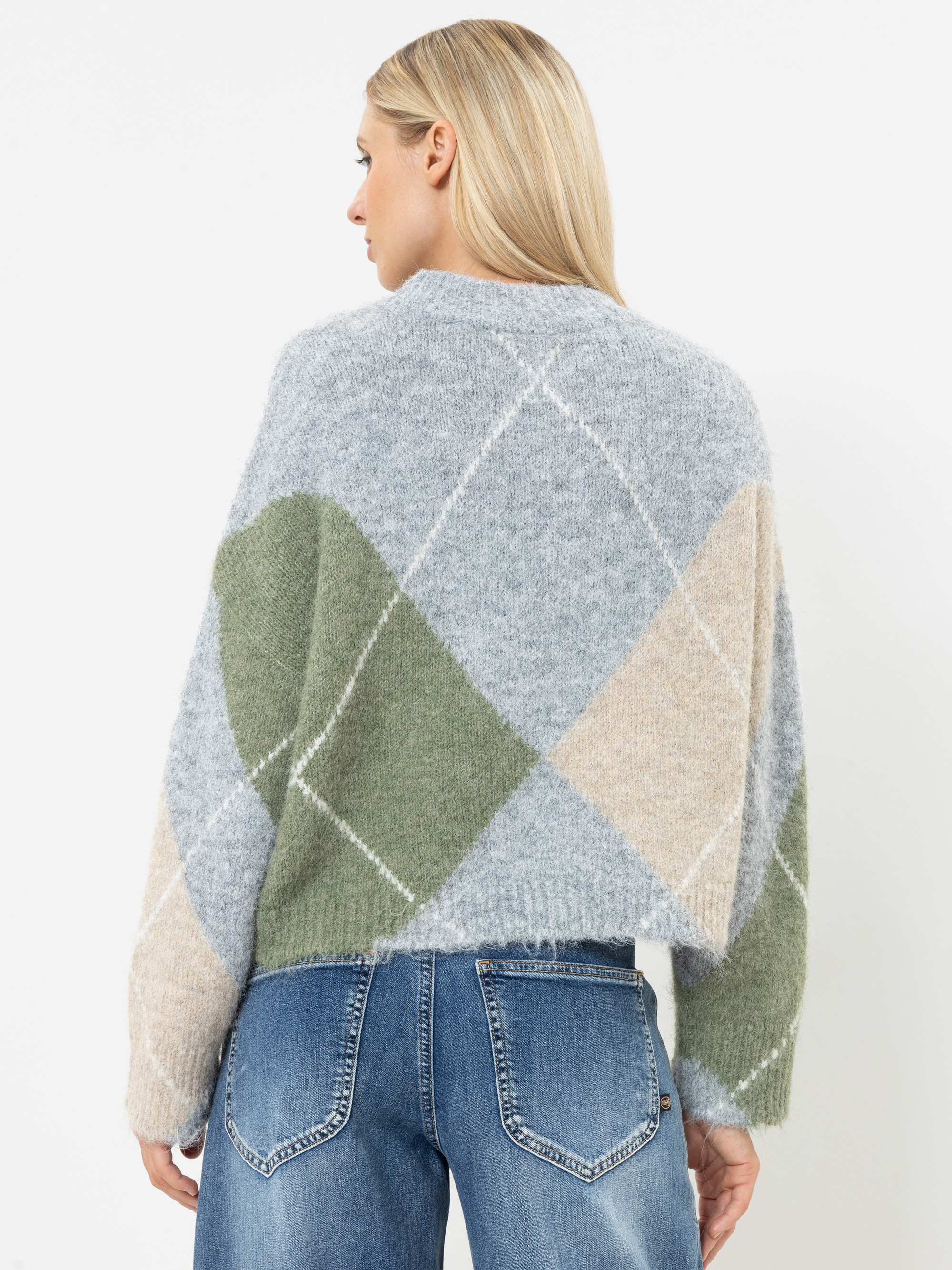 Eine Frau mit glattem blondem Haar trägt einen flauschigen, übergroßen Argyle-Pullover in Grau, Grün und Beige sowie eine blaue Jeans. Sie steht mit dem Rücken zur Kamera, sodass ihr Outfit vor einem schlichten weißen Hintergrund zu sehen ist.