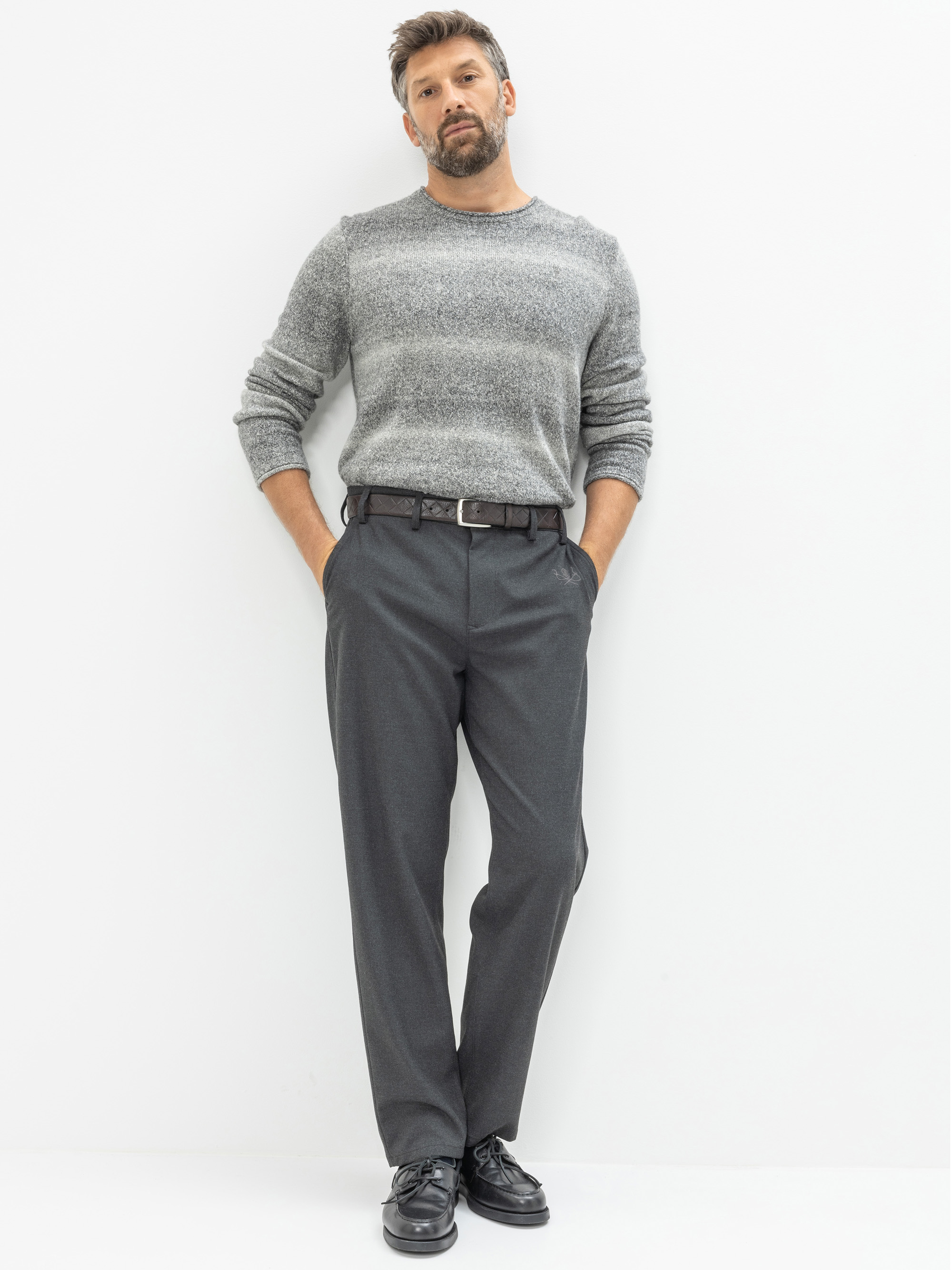 Ein Mann steht vor einem weißen Hintergrund und trägt einen grauen Pullover, eine dunkelgraue Anzughose, einen schwarzen Gürtel und schwarze Schuhe. Er hat die Hände in den Taschen und trägt kurzes Haar und einen Bart.