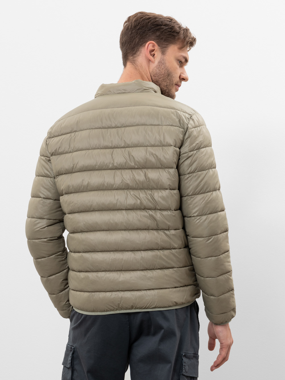 Ein Mann mit kurzen braunen Haaren blickt von sich weg, trägt eine hellolivgrüne Pufferjacke und eine dunkelgraue Cargohose und steht vor einem schlichten weißen Hintergrund.