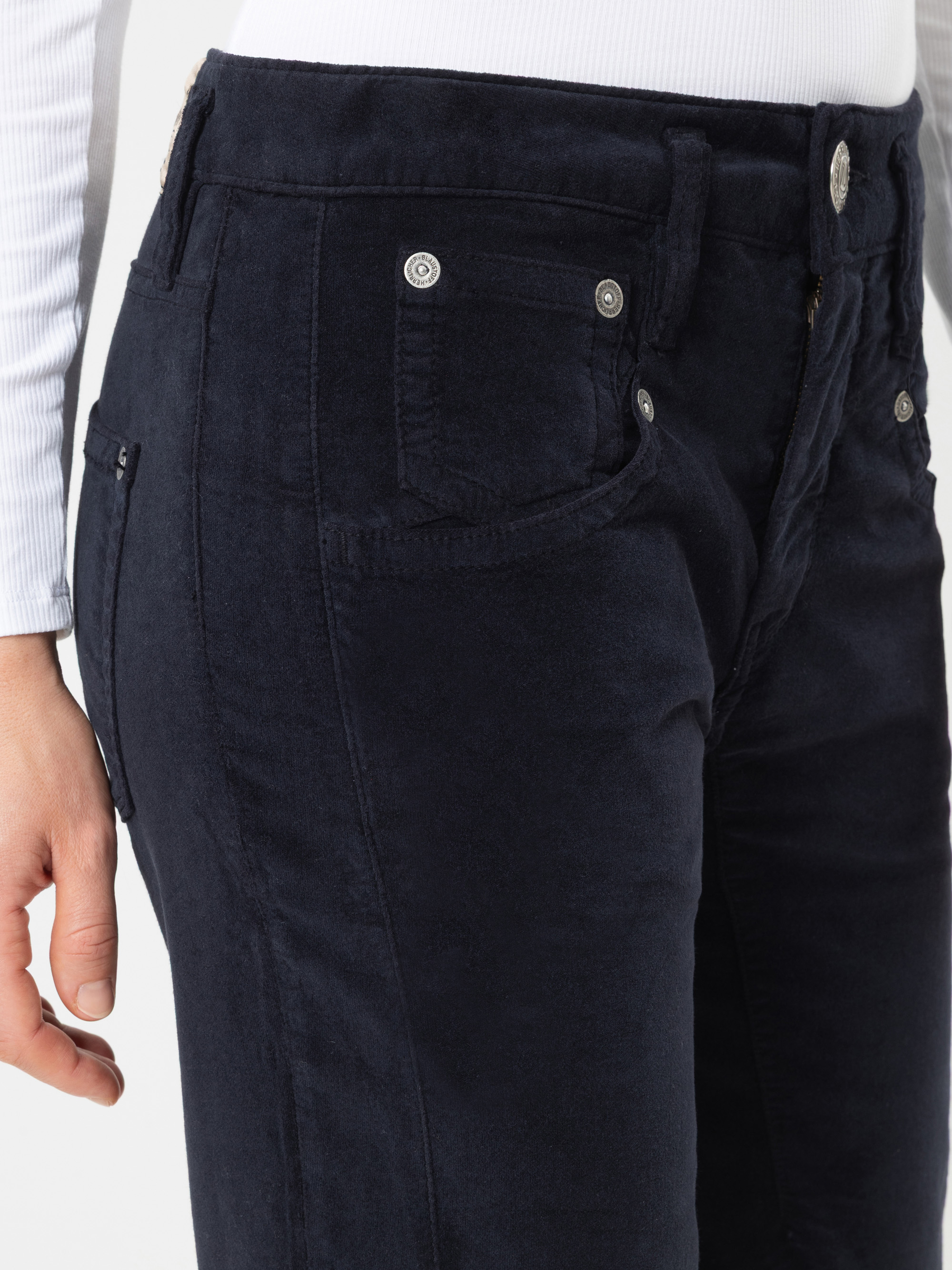 Eine Person, die eine dunkelblaue Hose mit hoher Taille und ein weißes, langärmeliges, geripptes Oberteil trägt. Der Fokus des Bildes liegt auf dem Bereich der linken Hüfte und der Taille; dabei sind die Vordertaschen der Hose und die Metallknöpfe zu