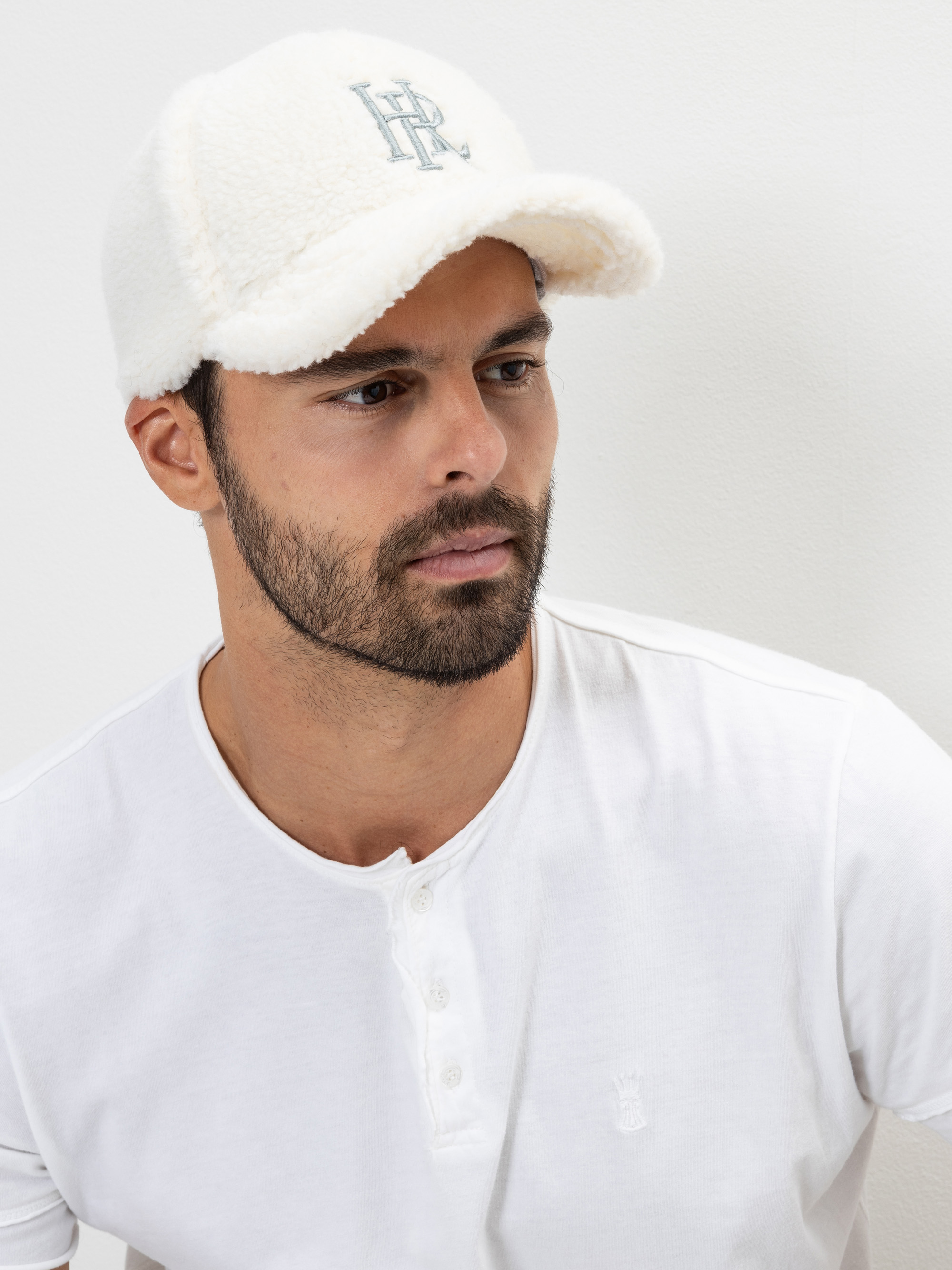 Ein Mann mit gestutztem Bart, der eine weiße Baseballmütze aus Shearling mit aufgestickten Initialen und ein weißes Henley-Shirt trägt und vor einem einfarbigen hellen Hintergrund leicht zur Seite schaut.