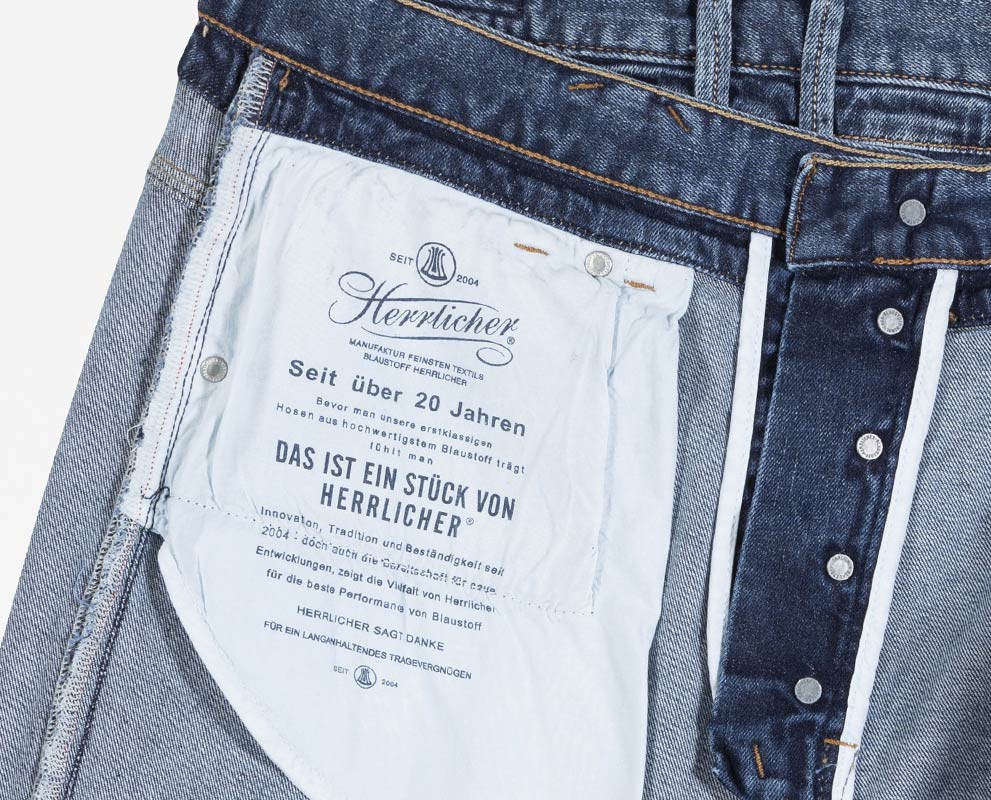 Nahaufnahme der Innenseite einer Jeans, die das Taschenfutter mit aufgedrucktem deutschen Text und dem Herrlicher-Markenlogo zeigt. Die Jeans ist aufgeknöpft, so dass die Metallknöpfe des Hosenschlitzes und der Denim-Stoff sichtbar werden.