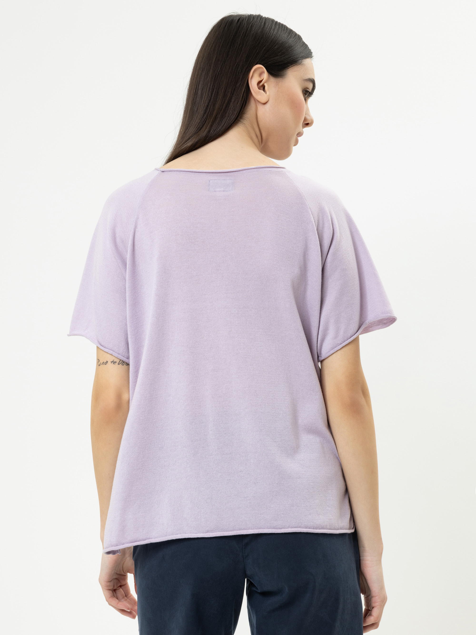 Eine Frau mit glattem dunklem Haar steht mit dem Rücken zur Kamera, trägt ein locker sitzendes, kurzärmeliges lavendelfarbenes Oberteil und eine dunkle Hose vor einem schlichten weißen Hintergrund.