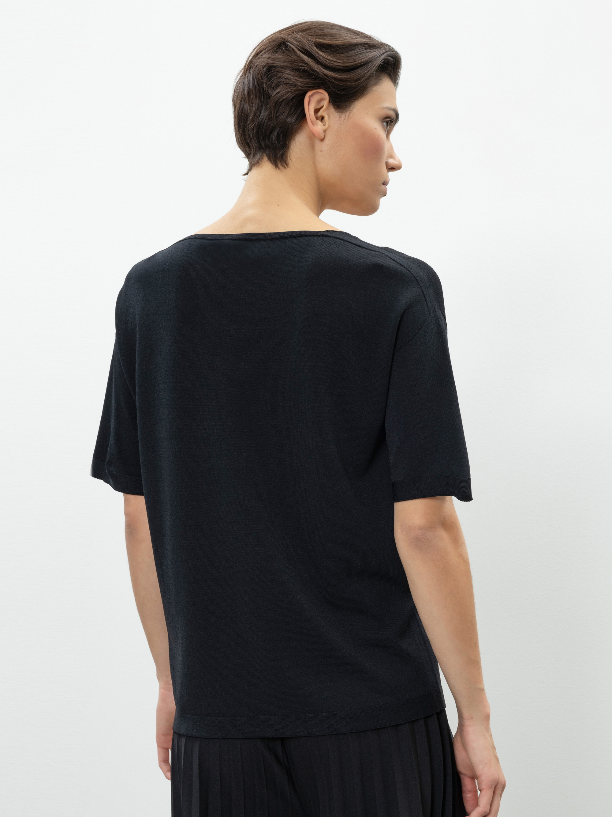 Eine Person mit kurzen braunen Haaren wird von hinten gezeigt, die ein locker sitzendes schwarzes kurzärmeliges Oberteil und eine schwarze Bundfaltenhose trägt und vor einem schlichten weißen Hintergrund steht.