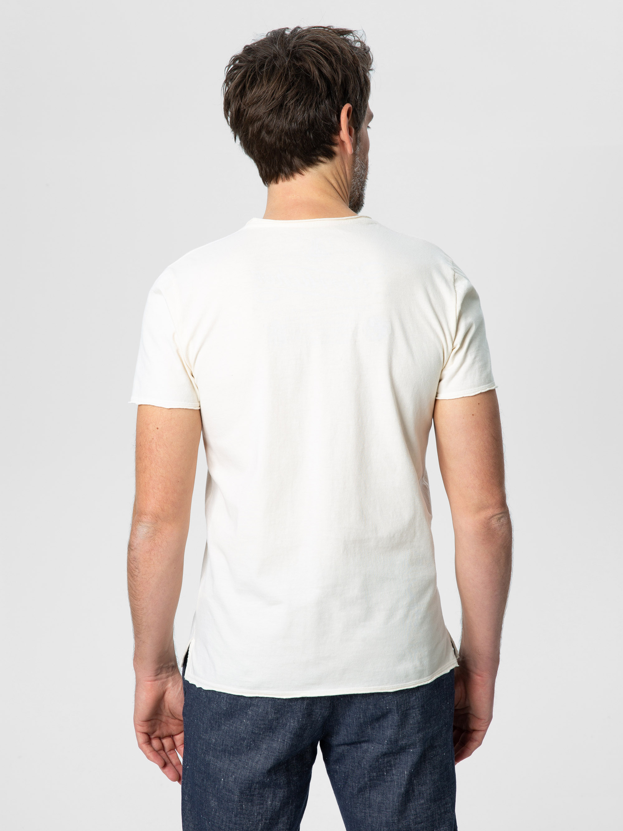 Ein Mann mit kurzen braunen Haaren steht mit dem Rücken zur Kamera, trägt ein schlichtes weißes kurzärmeliges T-Shirt und blaue Jeans vor einem hellgrauen Hintergrund.