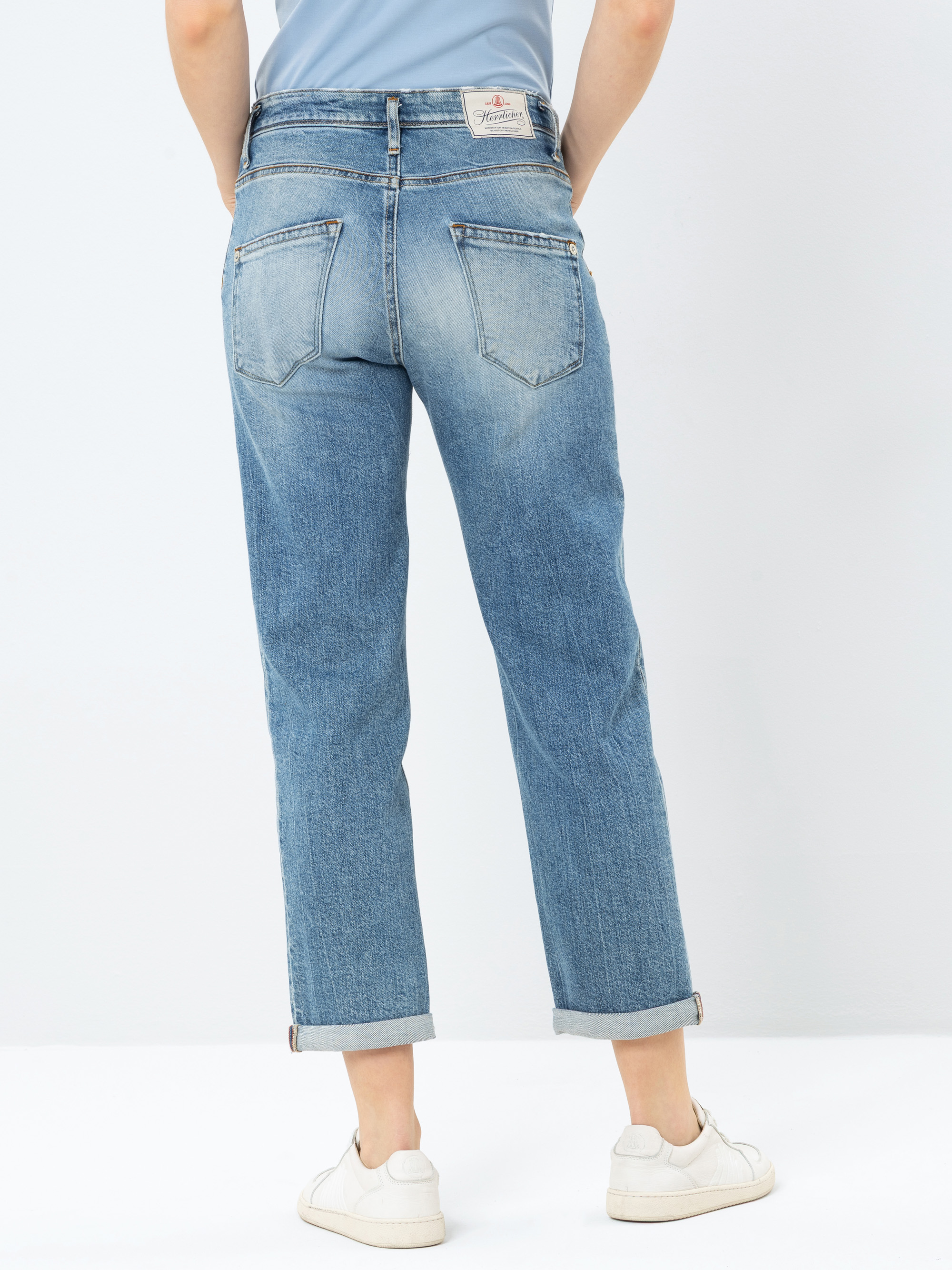 Person in hellblauer Jeans mit geradem Bein und weißen Turnschuhen, die mit dem Rücken zur Kamera vor einem schlichten weißen Hintergrund steht. Die Jeans hat zwei Gesäßtaschen und ein Markenetikett am Bund.