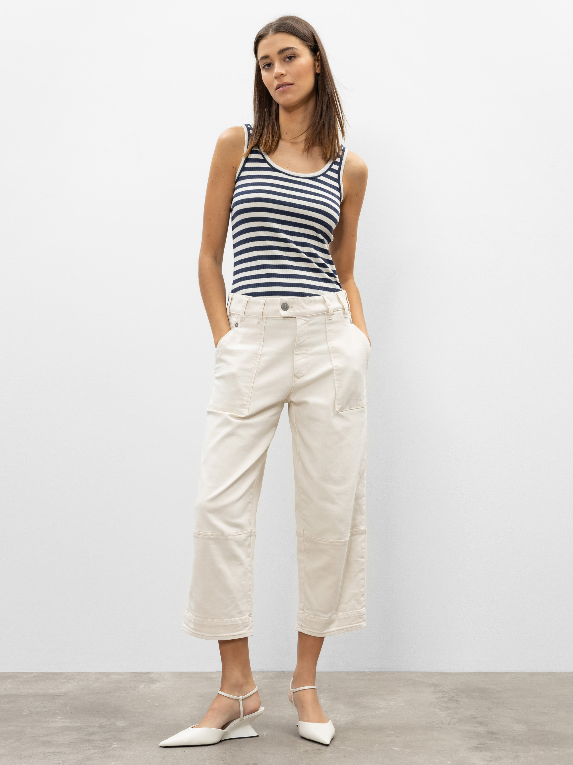 Eine Frau steht vor einem schlichten weißen Hintergrund, trägt ein marineblau-weiß gestreiftes Tanktop, eine cremefarbene Hose mit weitem Bein und weiße, spitze Schuhe, die Hände in den Taschen.
