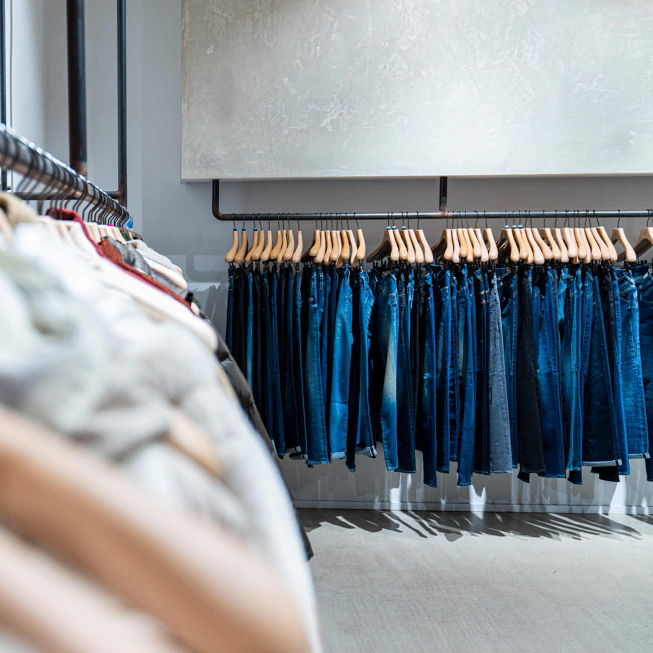 In einem hell erleuchteten Bekleidungsgeschäft hängt eine Reihe blauer Jeans auf hölzernen Bügeln an einem Metallgestell. Unscharfe Hemden oder Jacken sind im Vordergrund auf einem anderen Regal zu sehen.