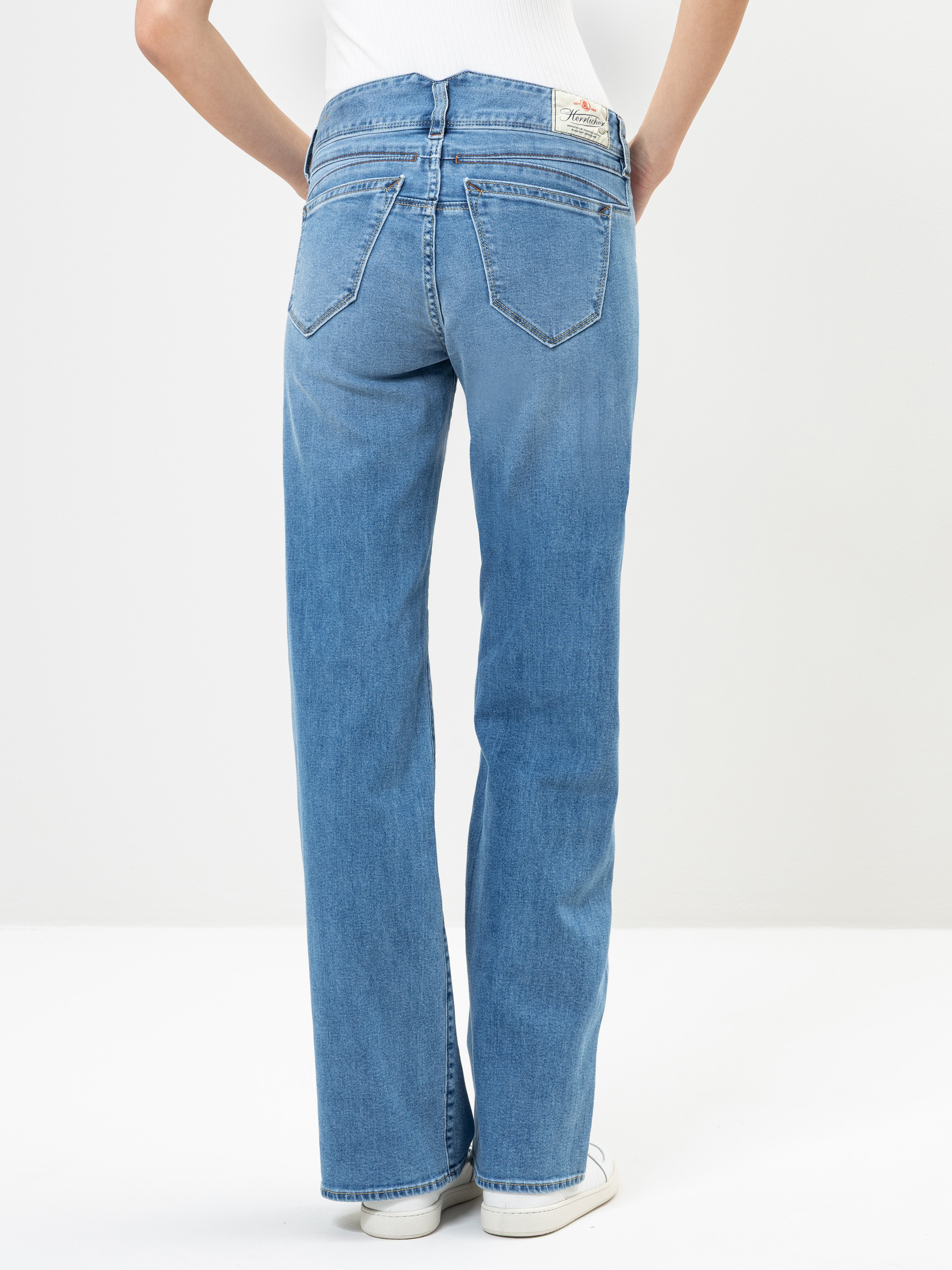 Person in hellblauer Jeans mit geradem Bein und weißen Turnschuhen, von hinten vor einem schlichten weißen Hintergrund dargestellt. Die Jeans hat zwei Gesäßtaschen und ein sichtbares Etikett am Bund.