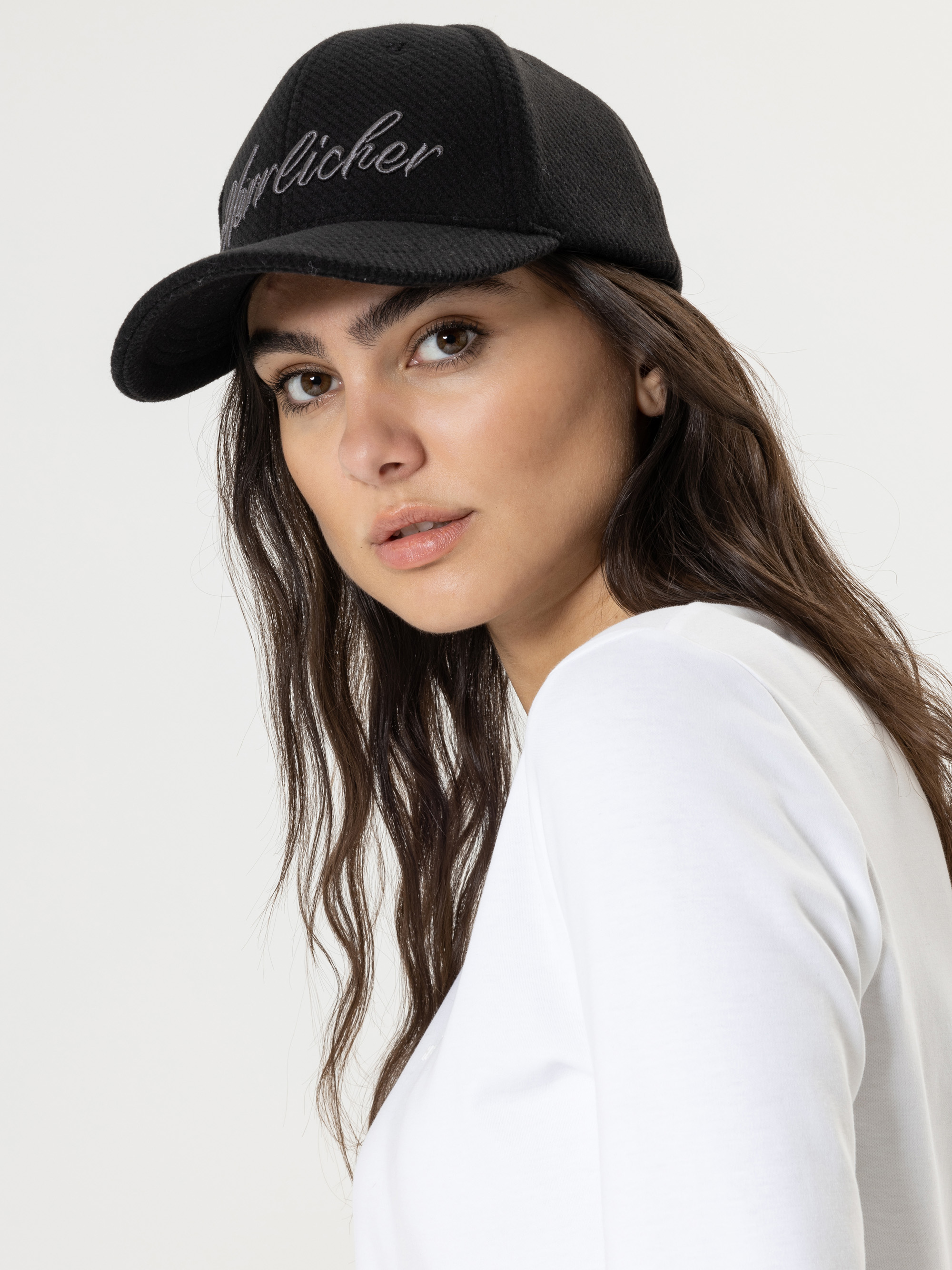 Eine Frau mit langen braunen Haaren, die eine schwarze Baseballkappe und ein weißes Hemd trägt, blickt vor einem schlichten hellen Hintergrund über ihre Schulter in die Kamera.
