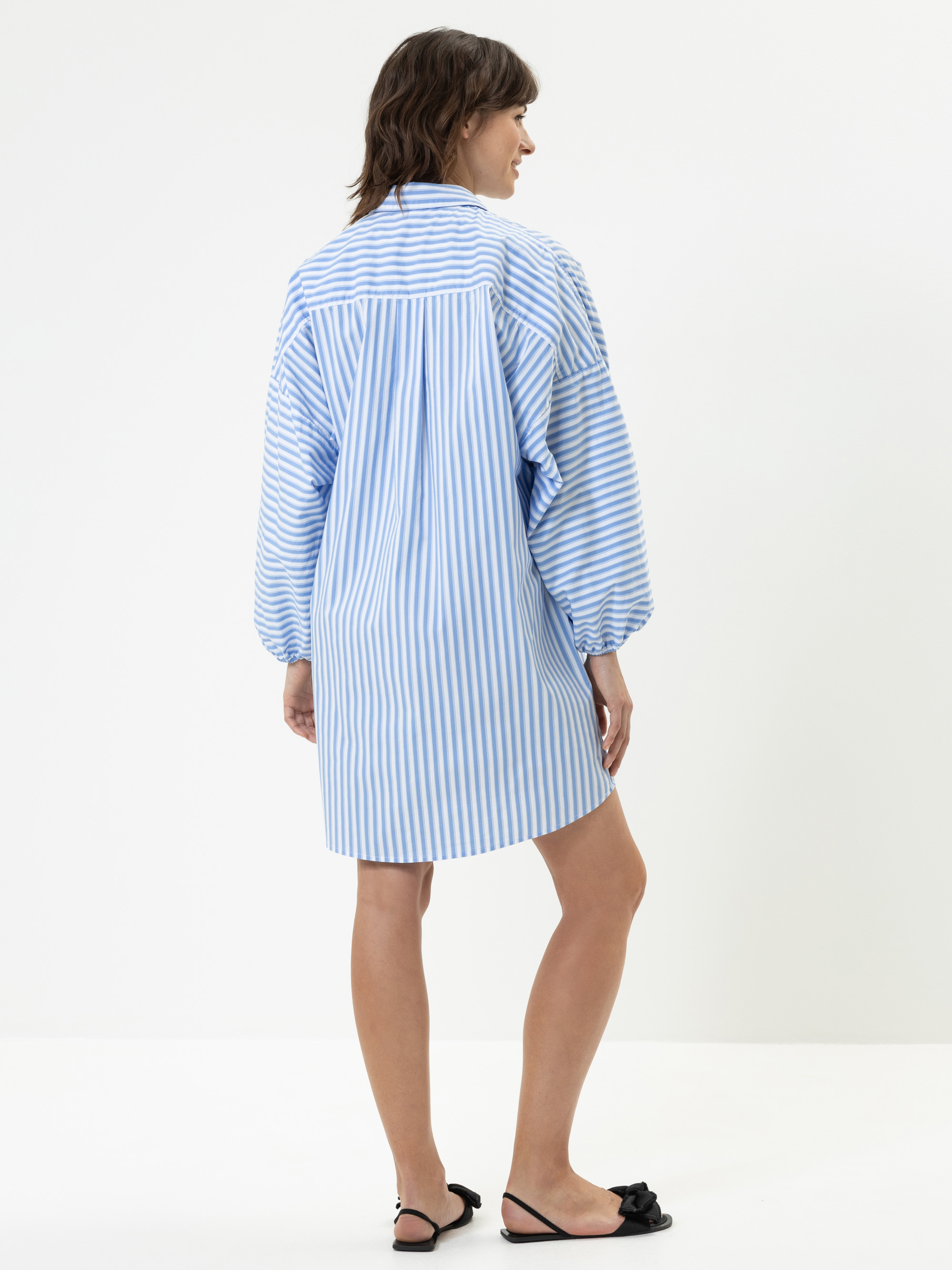 Eine Frau steht in einem lockeren, blau-weiß gestreiften, langärmeligen Hemdkleid und schwarzen Slippern vor einem schlichten weißen Hintergrund.
