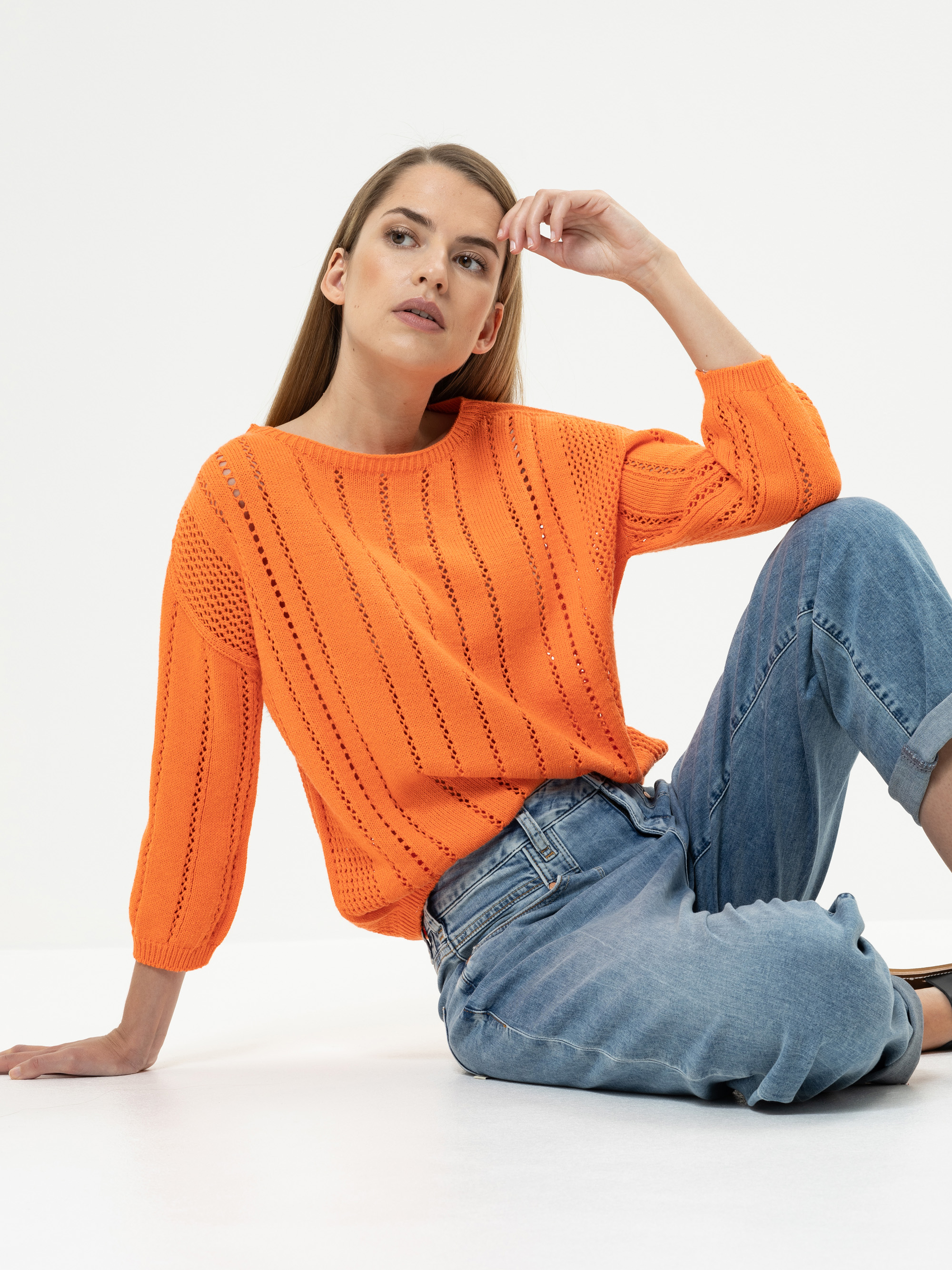 Eine Frau mit langen glatten Haaren sitzt auf dem Boden vor einem weißen Hintergrund. Sie trägt einen leuchtend orangefarbenen Strickpullover mit Dreiviertelärmeln und lässige Bluejeans und schaut mit nachdenklichem Blick leicht nach oben.